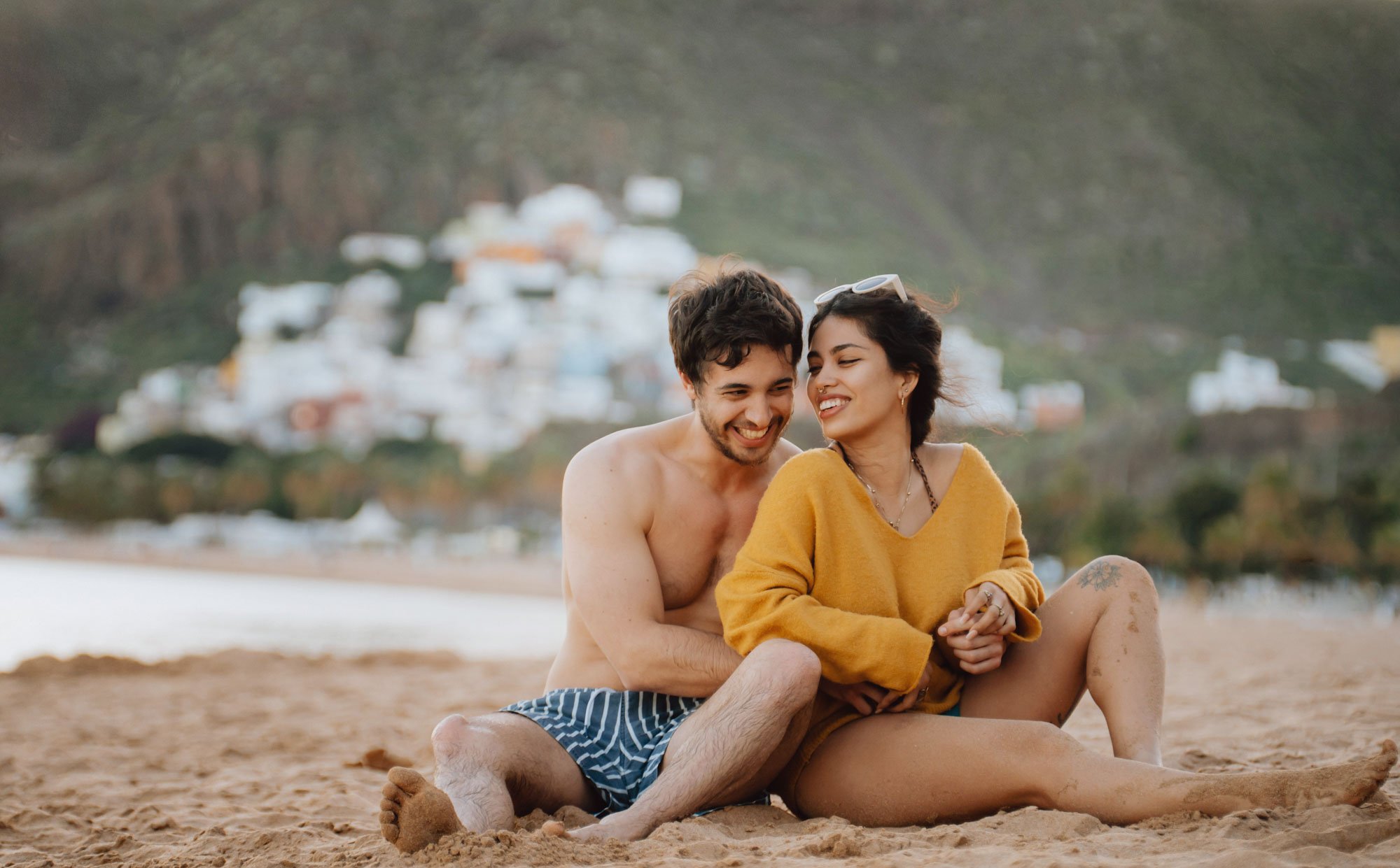 A young couple sitting closely on a sandy beach, smiling and enjoying each other's company, with a hillside town visible in the background.