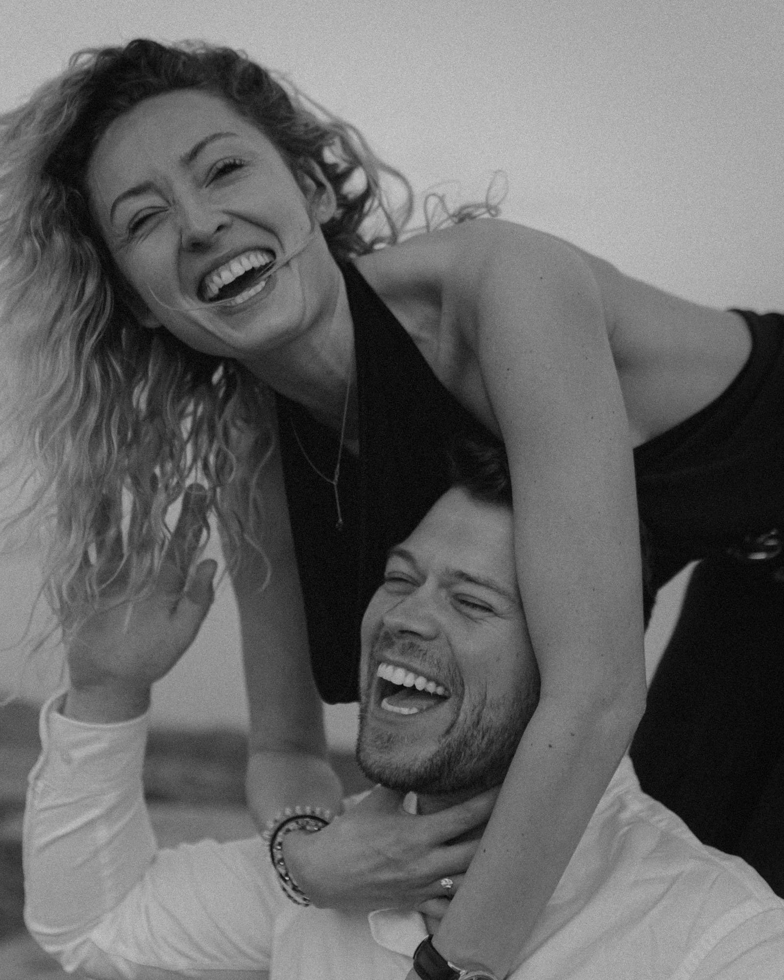 A woman with curly hair and a necklace playfully sitting on a man's shoulders, both laughing joyfully in a black and white photo. Alicante 