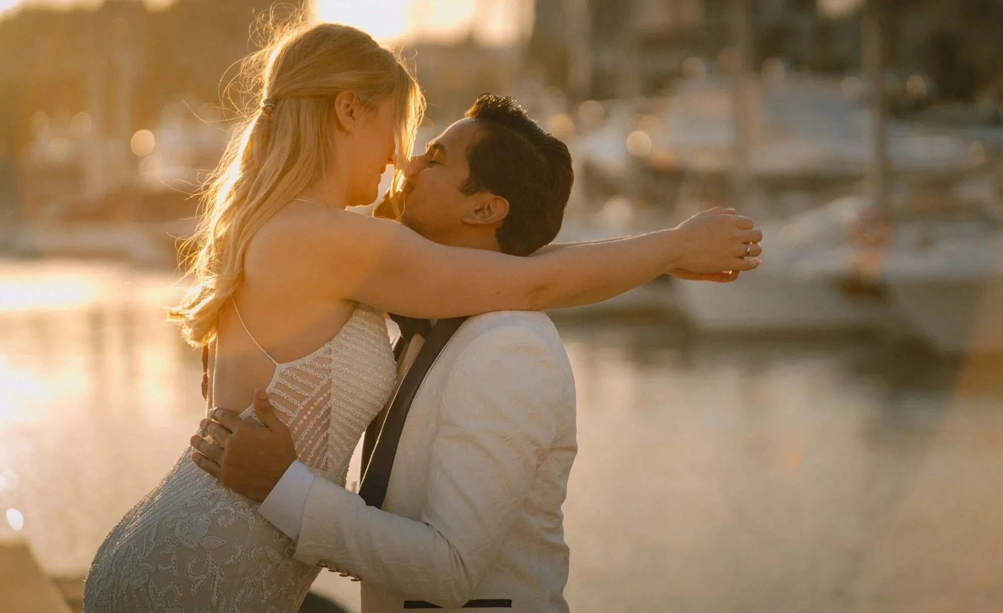 A couple embracing and kissing by a marina at sunset, with boats and water in the background.