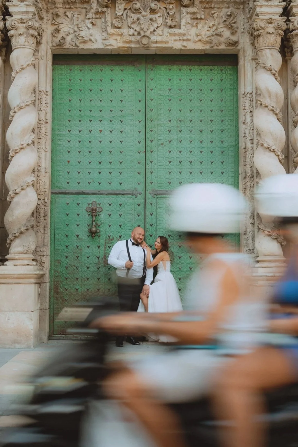 A couple in wedding attire standing in front of a large ornate green door with detailed stone carvings, with blurred cyclists passing by in the foreground.