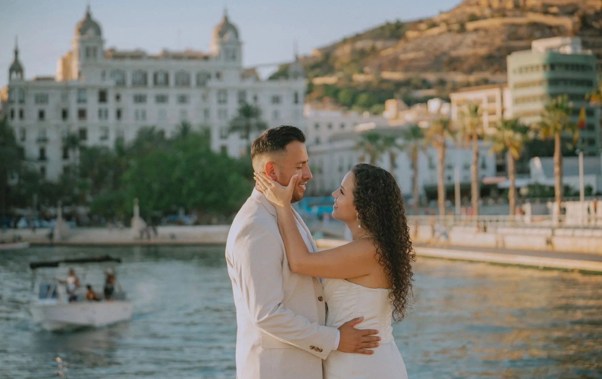 Couple in white wedding attire embracing by the water with boats, trees, city buildings, and hills in the background.