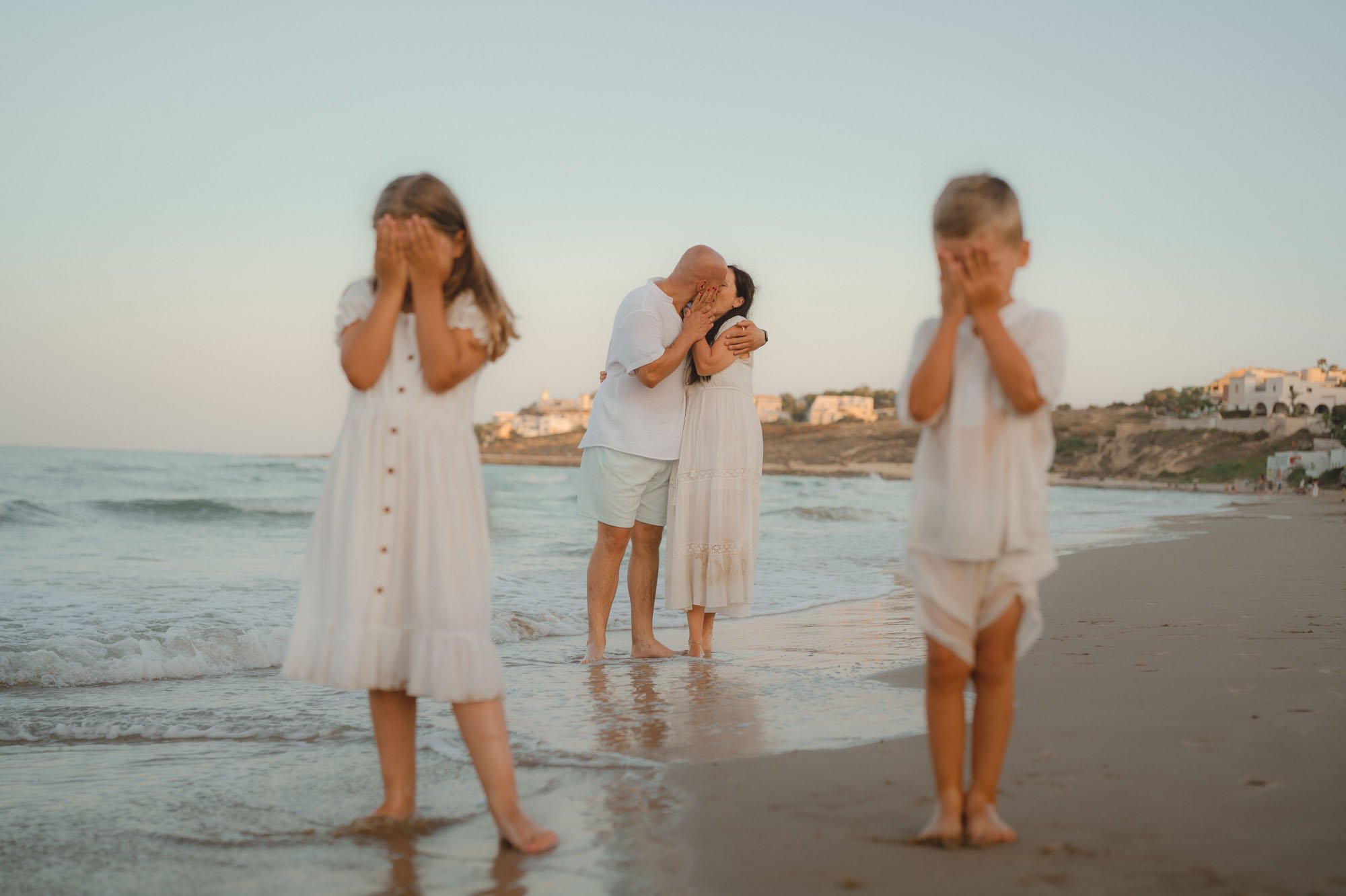 A family on a beach with a father and mother sharing a kiss in the background while two children, a girl and a boy, stand in the foreground with their hands covering their faces.