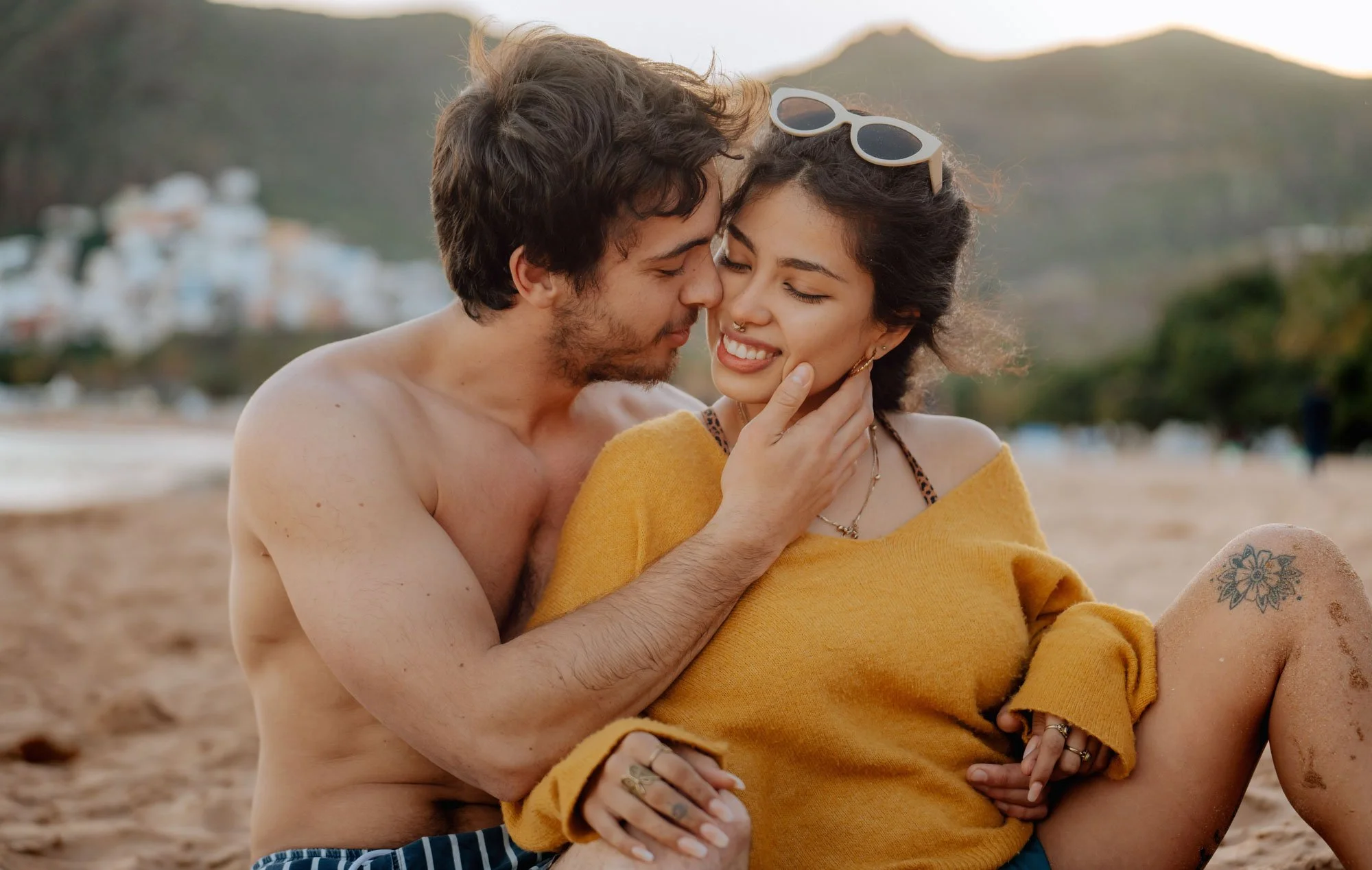A young couple sitting on the beach, smiling, with the man touching the woman's face. The woman is wearing sunglasses on her head, a yellow sweater, and has a tattoo on her thigh. In the background, there are hills and a town.