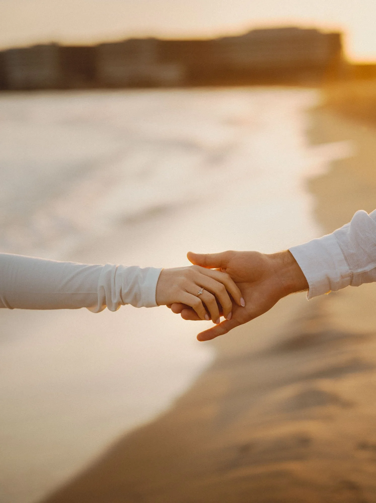 A close-up of two people holding hands; one person wearing a white long-sleeve shirt, the other wearing a beige or light tan sleeve. The background features a sandy beach and water at sunset.
