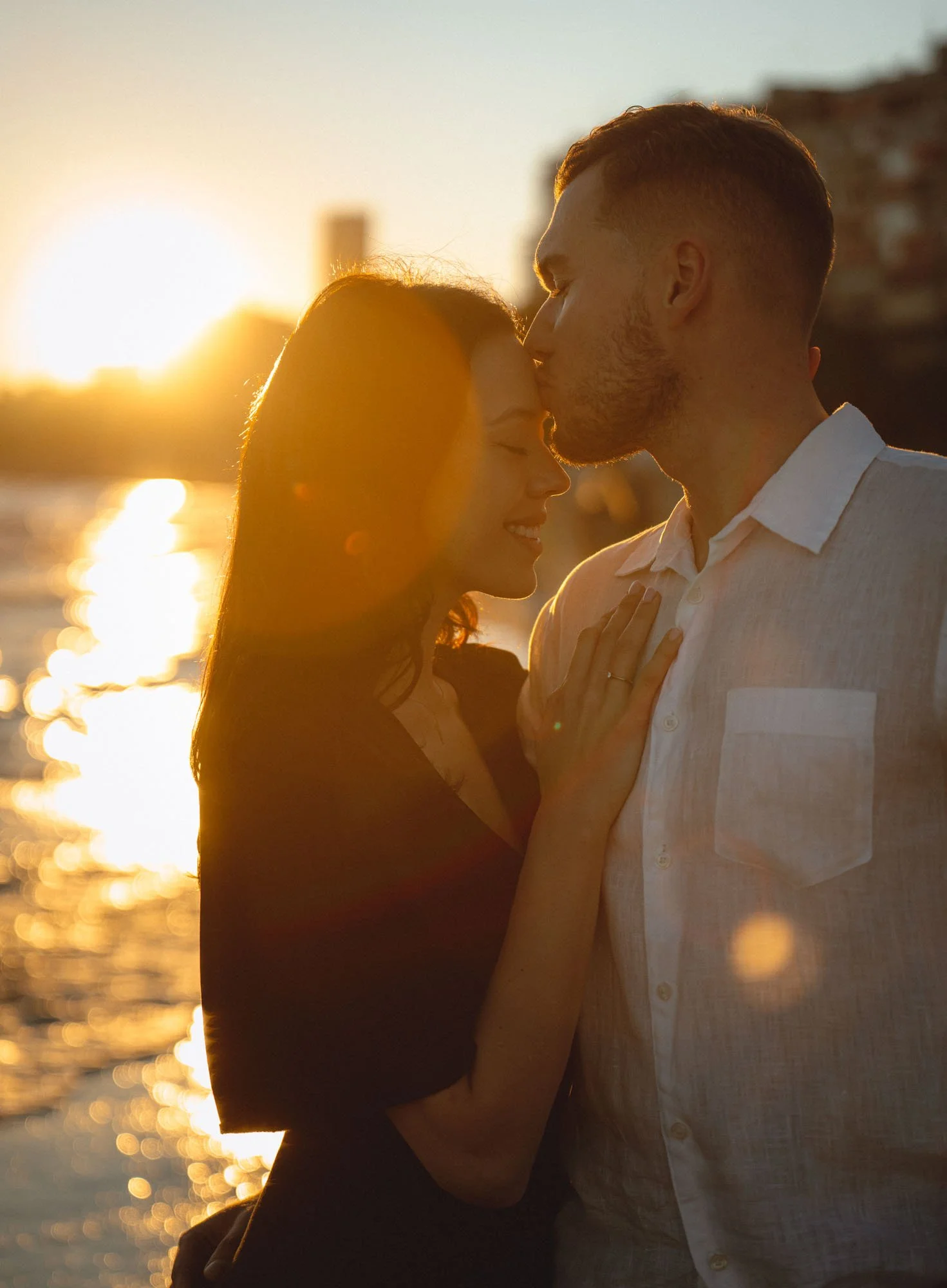 A couple stands close together at sunset, with the man kissing the woman's forehead, by the water.