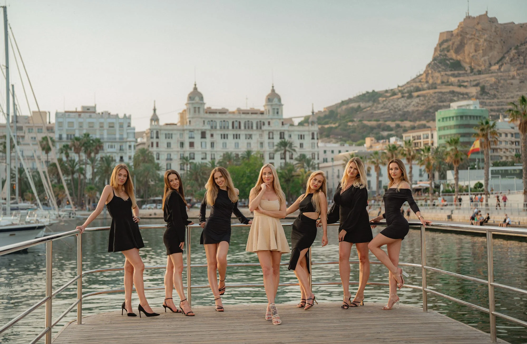 Group of seven women posing on a wooden dock by a marina with water, sailboats, and a cityscape with white buildings, palm trees, and a mountain in the background.