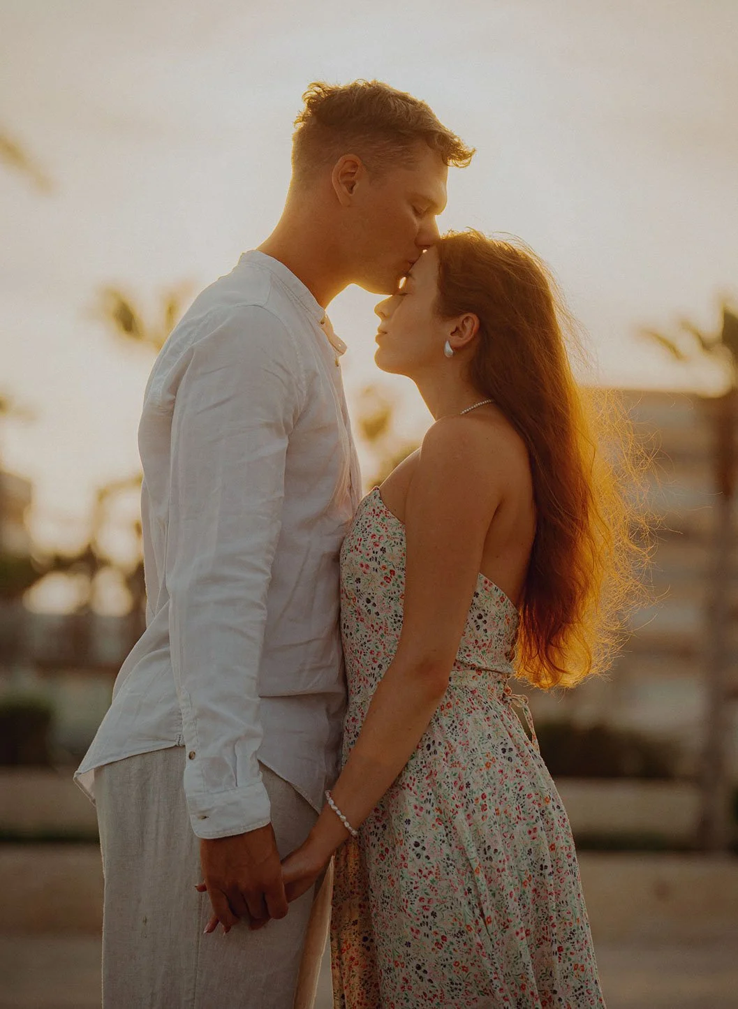 A couple standing outdoors at sunset, holding hands, with the man kissing the woman's forehead Alicante 