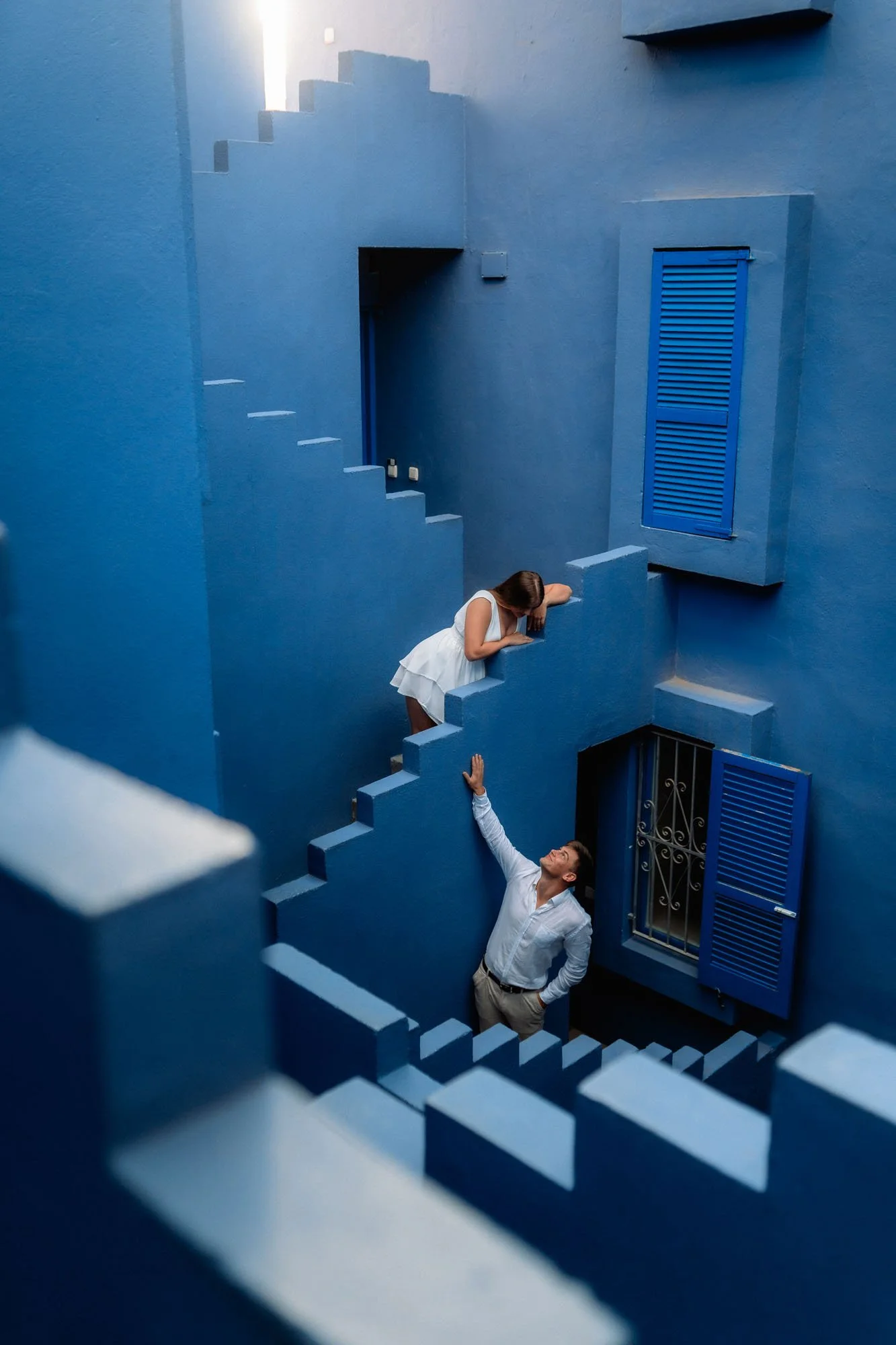 A woman in a white dress leaning over a blue balcony railing, reaching out to a man below, in a blue-walled courtyard with blue shutters. Calpe, Alicante Muralla Roja 