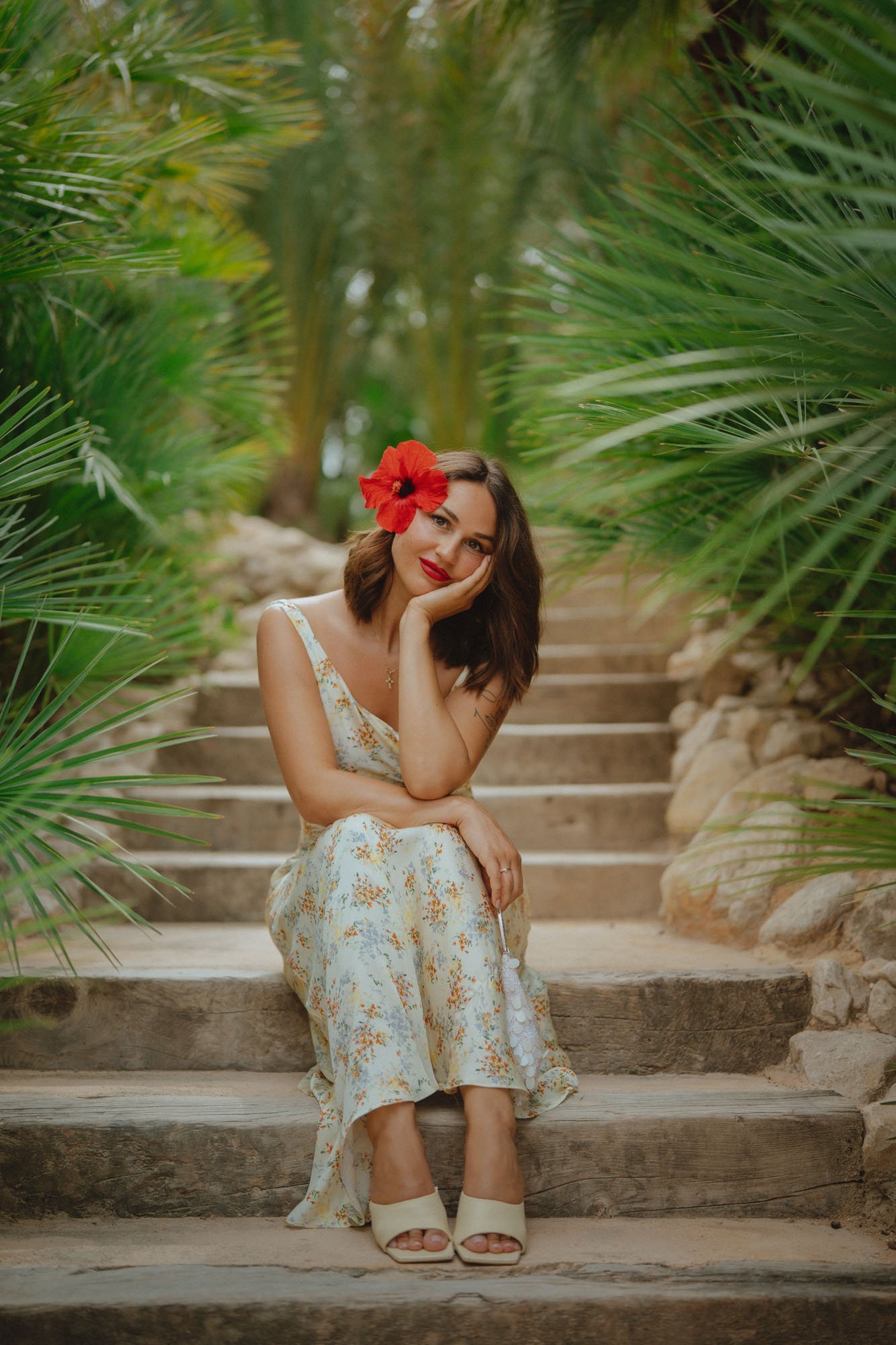 Woman sitting on wooden steps surrounded by lush green tropical plants, wearing a floral dress, with a red flower in her hair and beige high heels.