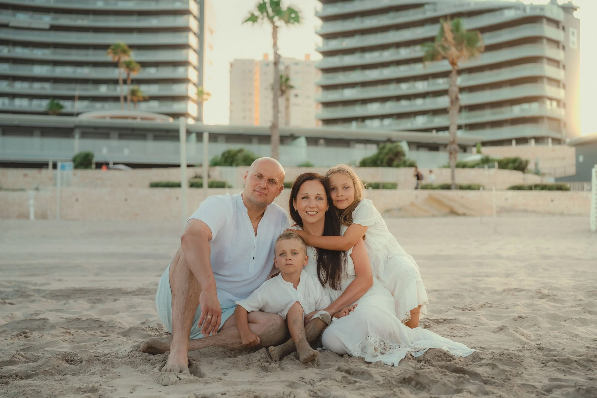A family of four sitting on the sand at the beach in front of tall buildings and palm trees during sunset, all dressed in white.