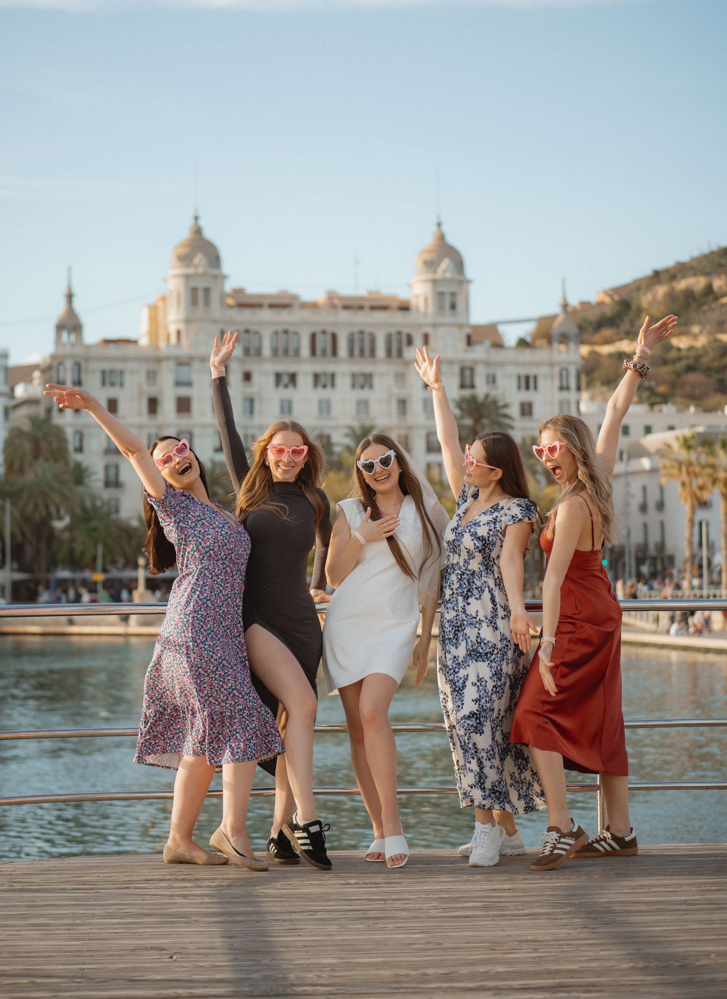Group of five women celebrating together on a pier, with a large historic building and a mountain in the background.