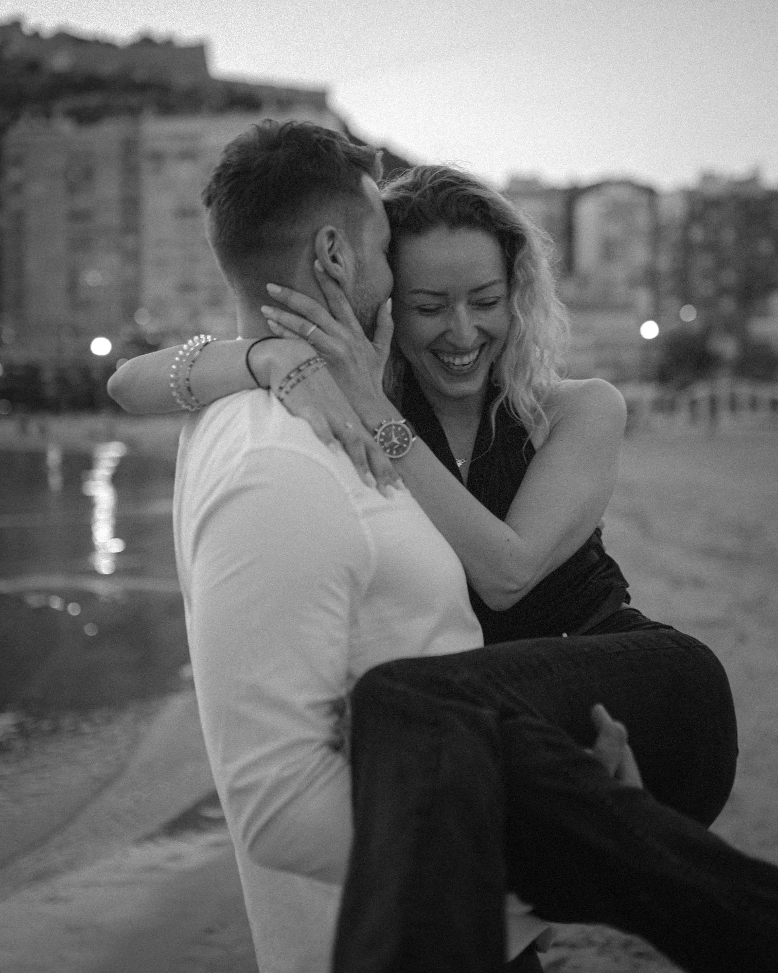 A man lifts a woman by the water, both smiling and engaged in a joyful moment at dusk or evening.