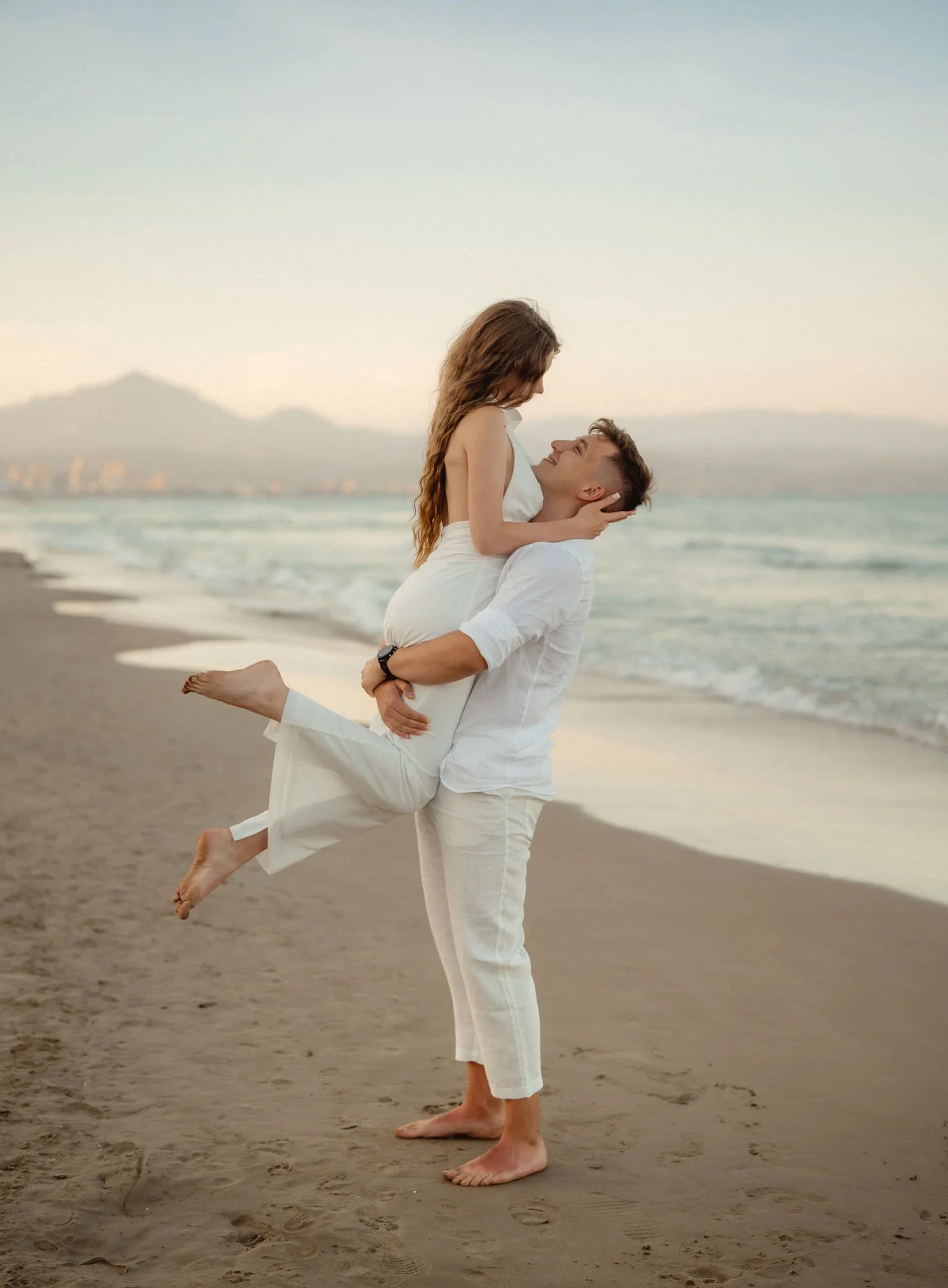A man lifting a woman at the beach during sunset, both wearing white clothing, with the ocean and distant mountains in the background.