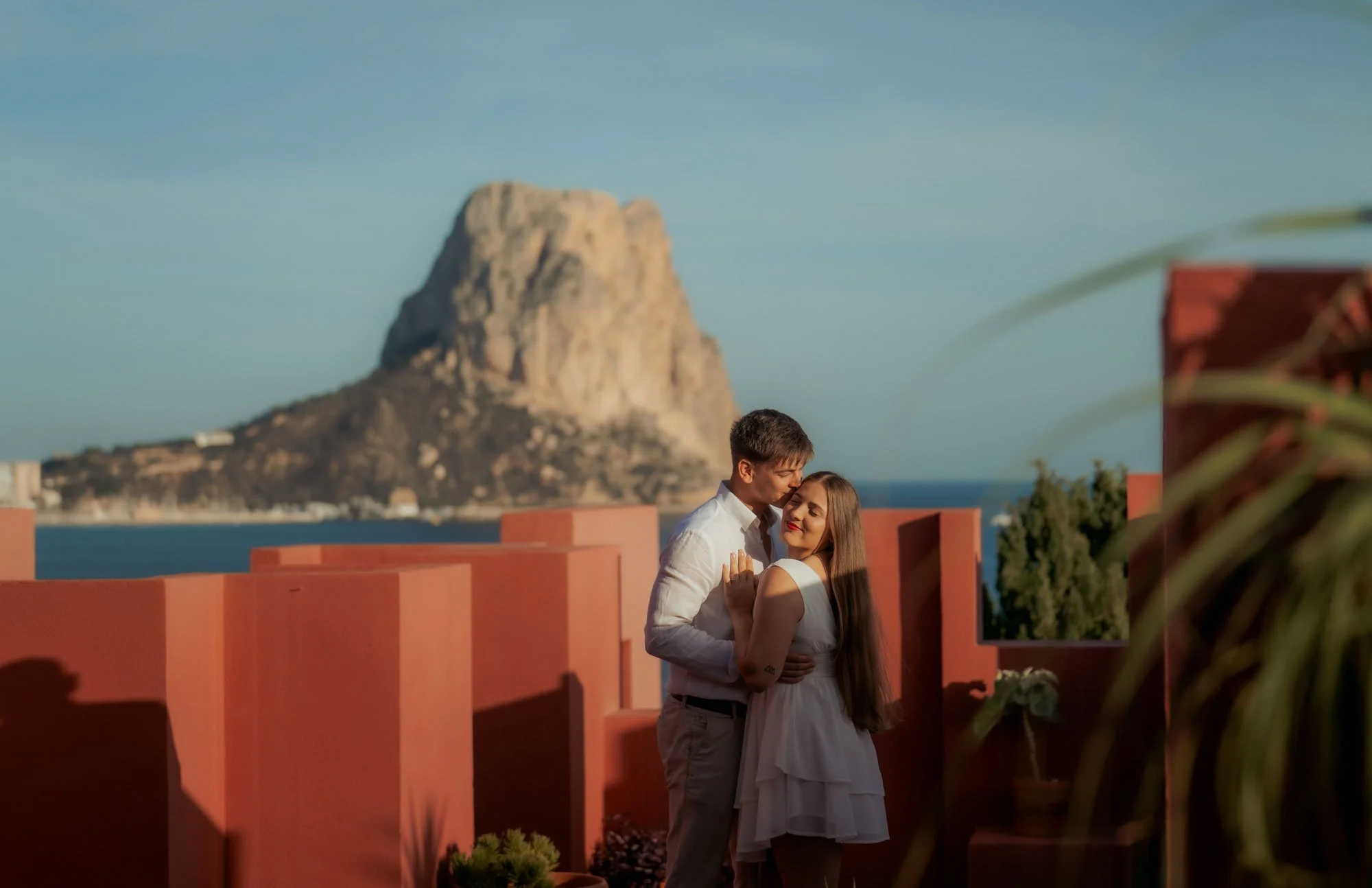 A couple embracing on a terrace with red walls, overlooking a body of water and a large mountain in the background during sunset.