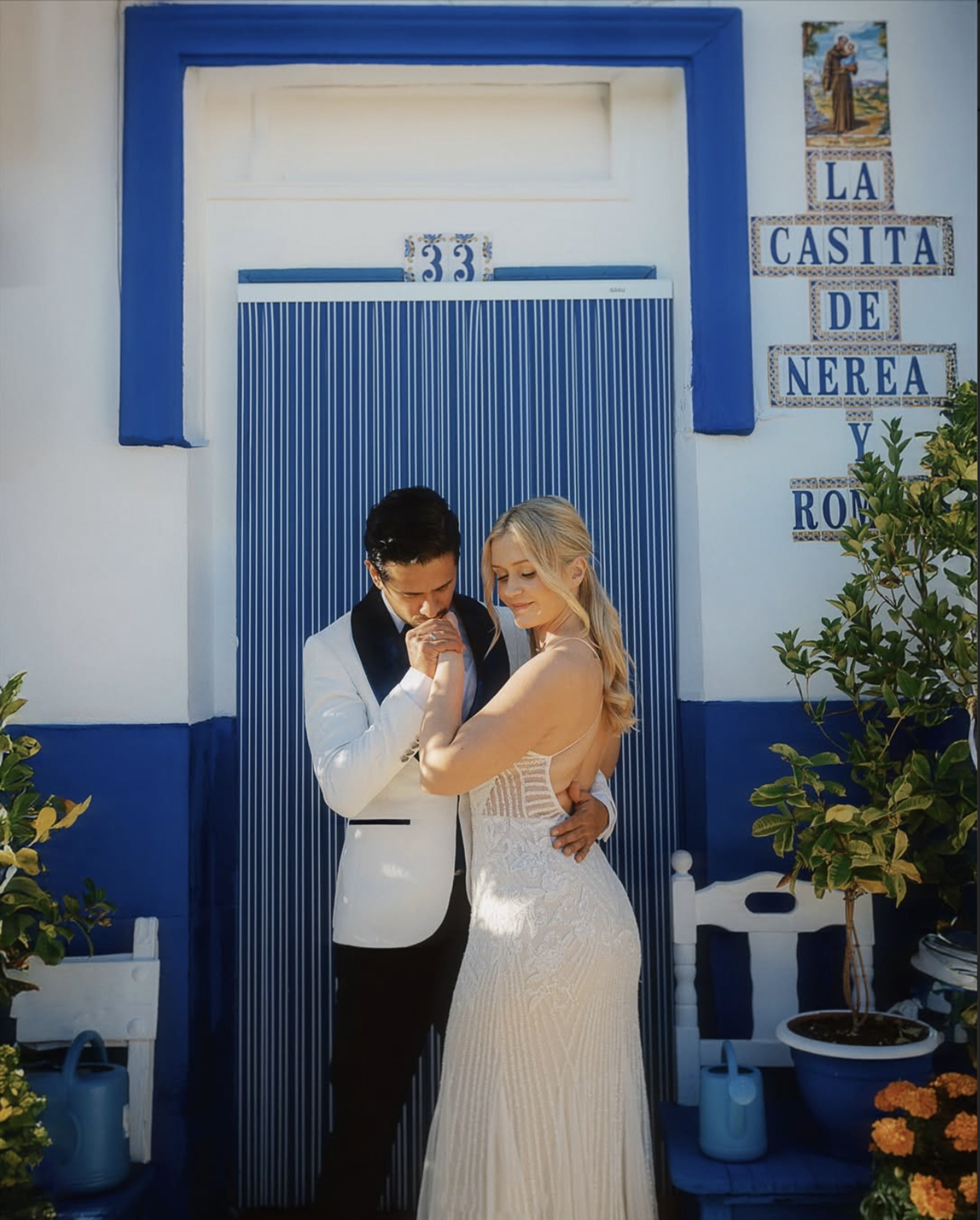 Para młoda pozują razem przed niebiesko-białymi drzwiami na weselu, na ścianie widnieje tabliczka z napisem „La Casita de Nerea y Roni”. Alicante Stare miasto, hiszpania