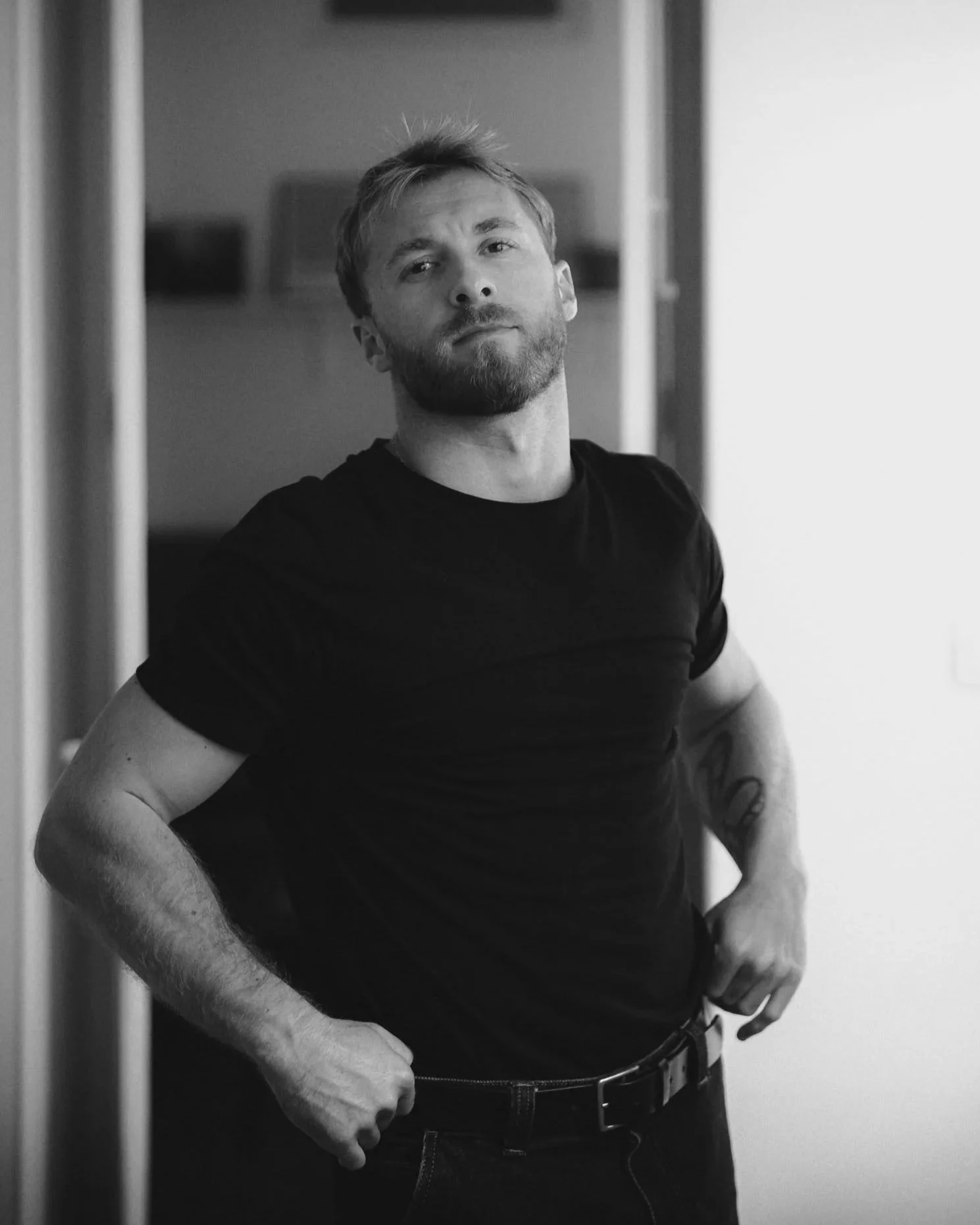 Czarno-białe zdjęcie mężczyzny z brodą i krótkimi włosami, ubranego w czarny T-shirt i dżinsy, stojącego z rękami pod boki i patrzącego w obiektyw. Fotograf Konrad Maziarz Alicante