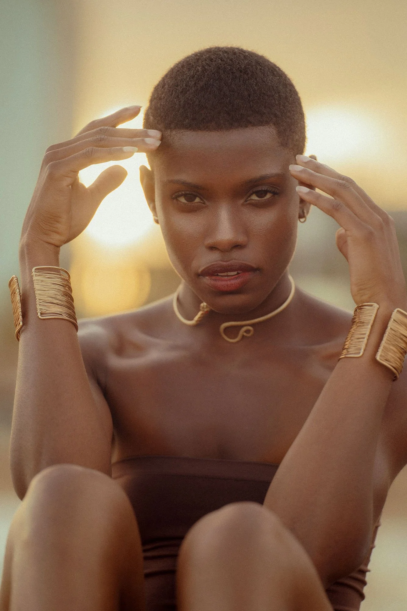 A young woman with short hair and dark skin, wearing gold jewelry, poses outdoors at sunset.