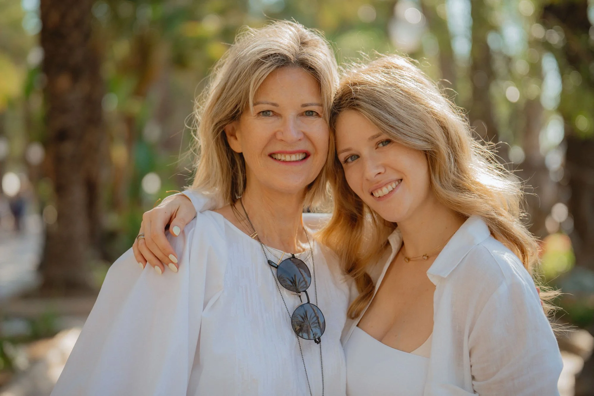 Two smiling women, one older and one younger, standing close outdoors with arms around each other.