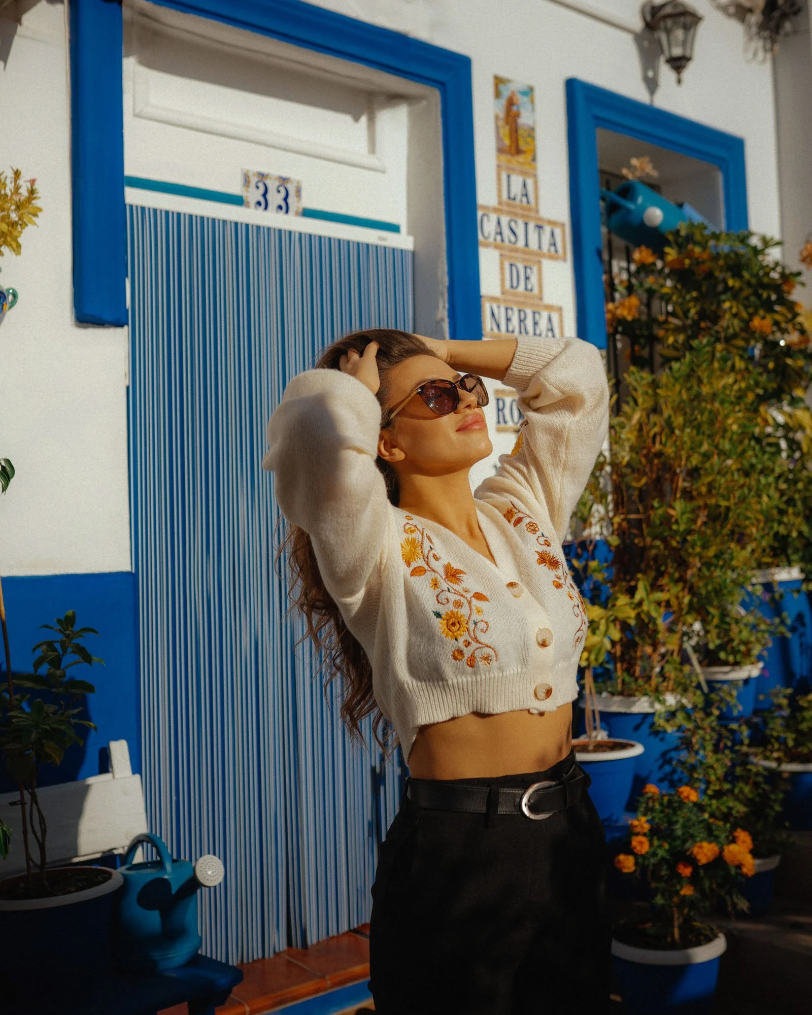 A woman with long hair wearing sunglasses, a cream-colored embroidered cropped cardigan, and black high-waisted pants with a belt, stands outdoors with her hands in her hair, enjoying the sunlight in front of a blue and white building with potted pla
