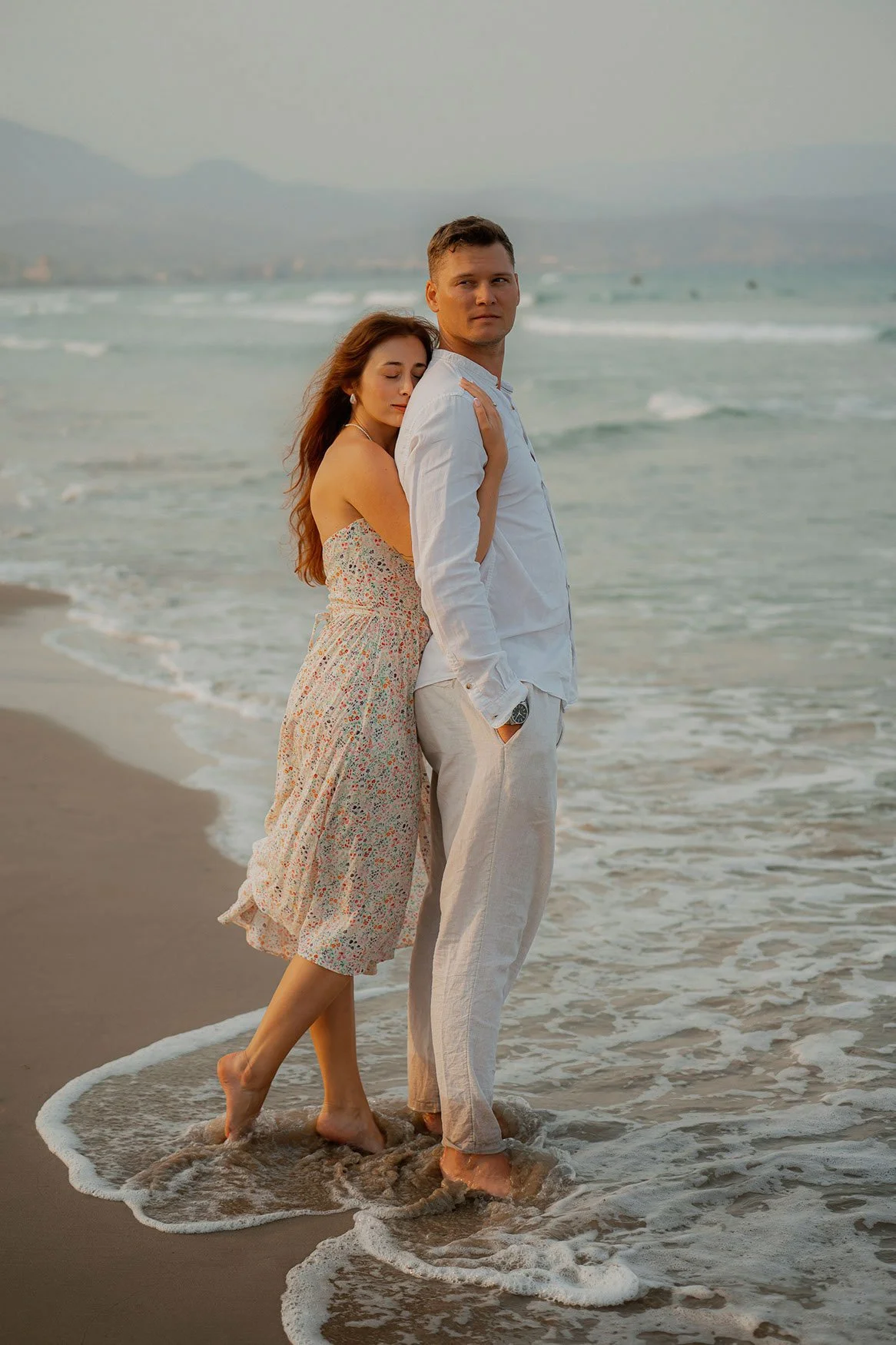 A couple standing close together in the shallow surf at the beach during sunset, with mountains and cloudy sky in the background.