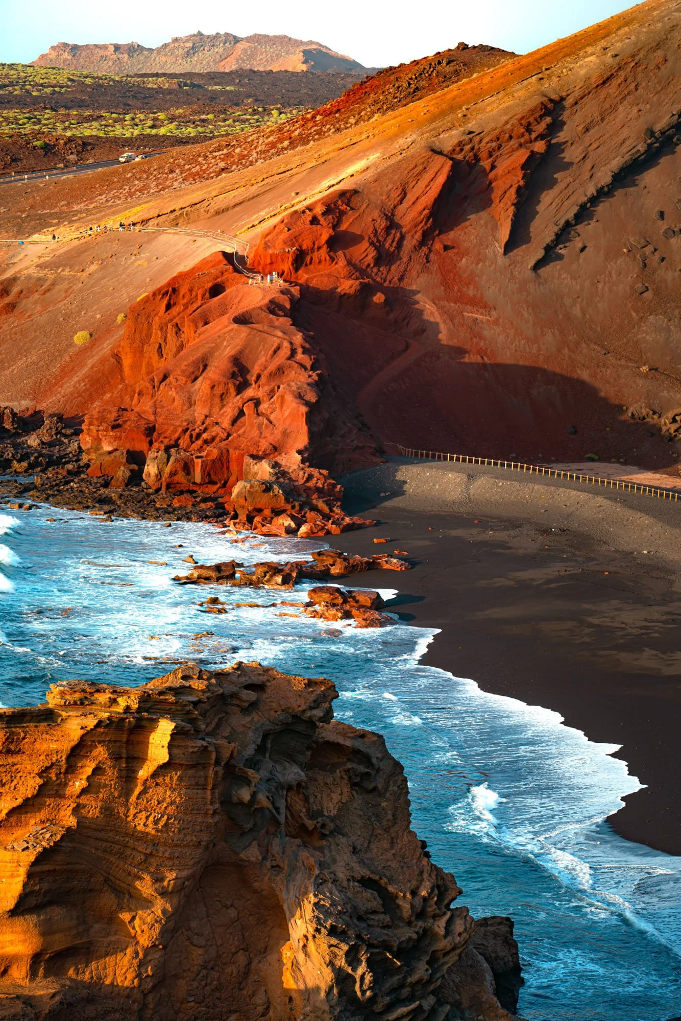 A rugged coastal landscape with red and orange rock formations, a black sand beach, and waves crashing against the rocks. In the background, there are hills and mountains under a clear sky.