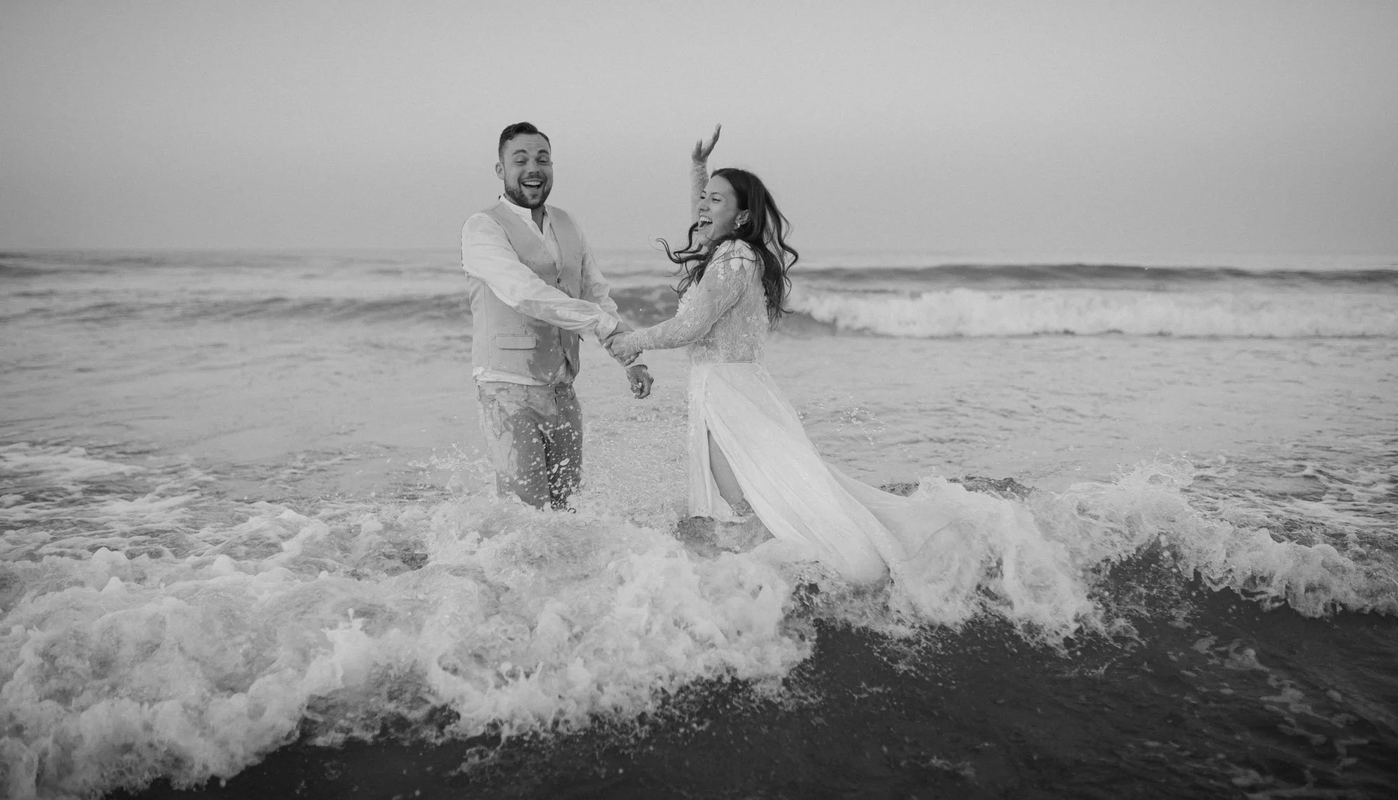 A joyful couple in wedding attire holding hands and playing in the ocean waves at the beach.