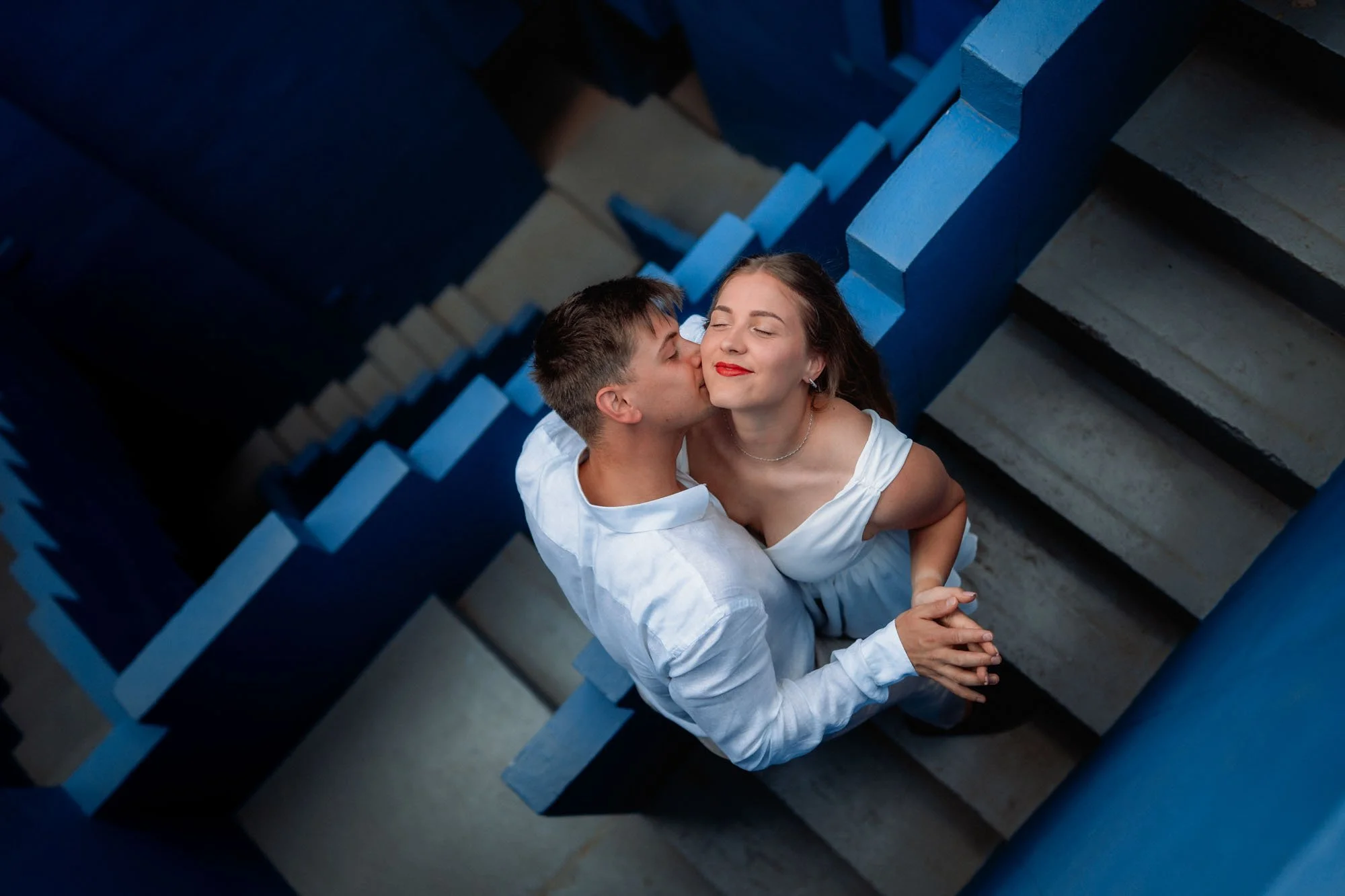 A couple standing on a staircase, with the man kissing the woman's cheek, both dressed in white, with the woman smiling with eyes closed, surrounded by blue and gray stair walls.