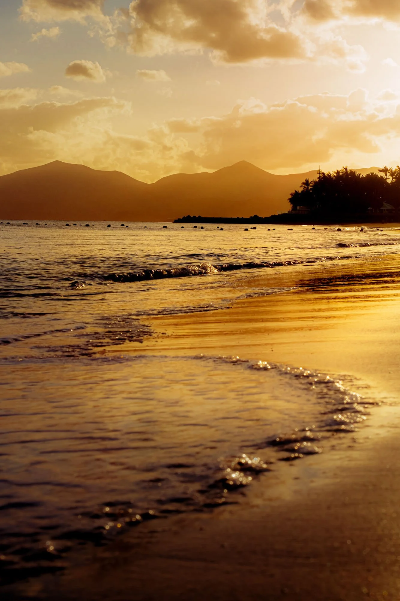 Sunset over the ocean with mountains in the background and waves gently lapping at the sandy beach in the foreground.