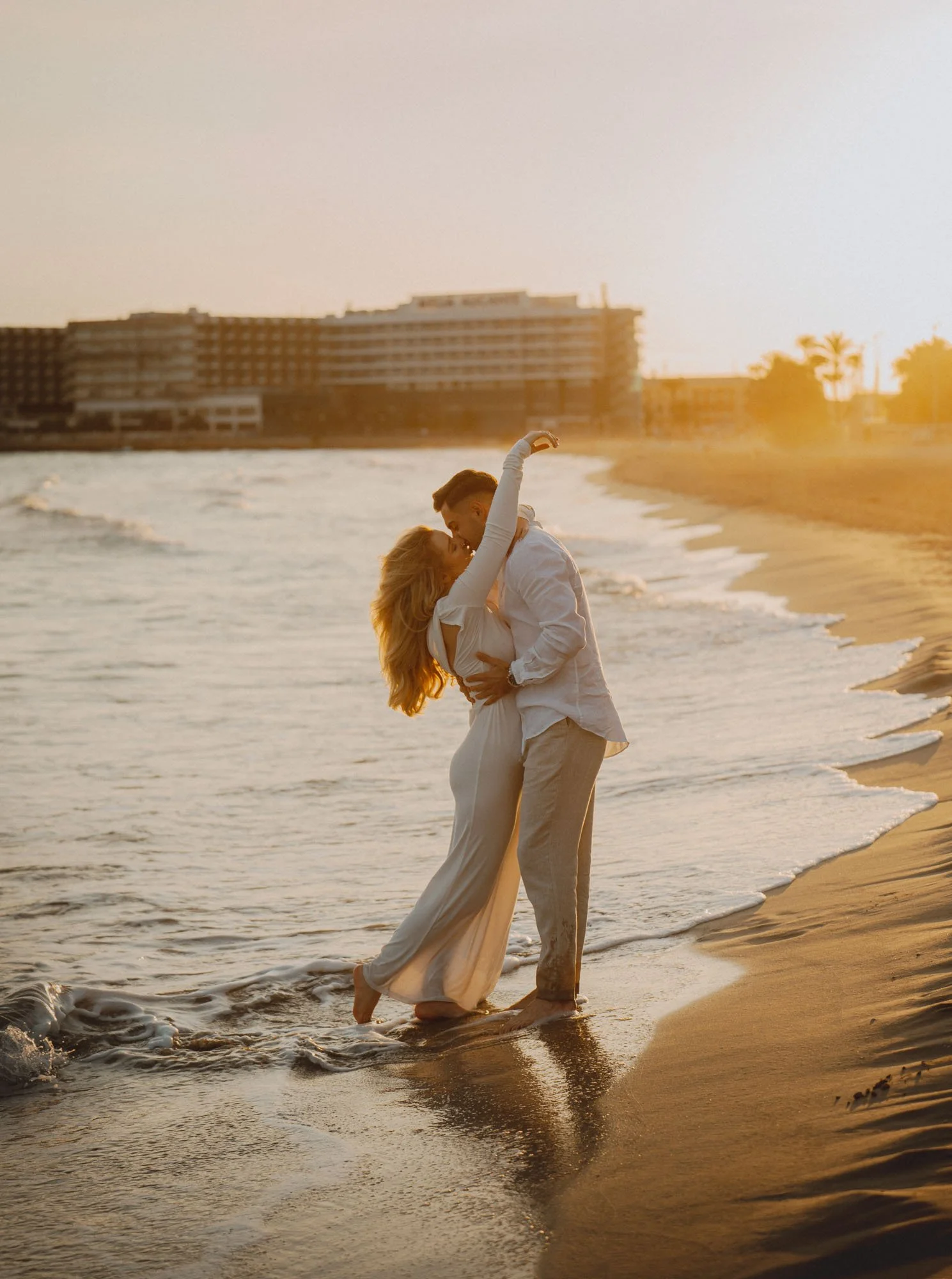 Para tańcząca i obejmująca się na plaży podczas zachodu słońca, na tle fal oceanu i panoramy miasta. Plaża Postiguet Alicante