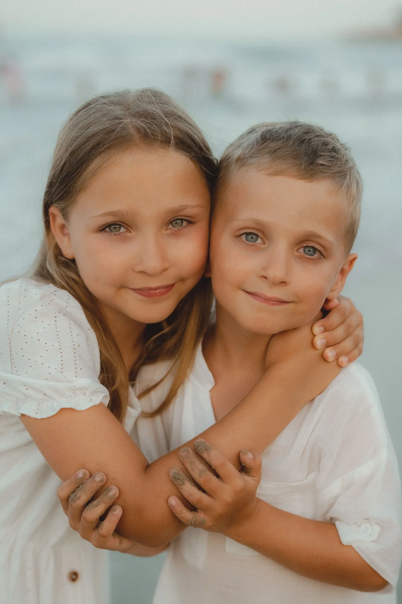 A girl and a boy hugging each other, both with sandy hands, standing outdoors near water.
