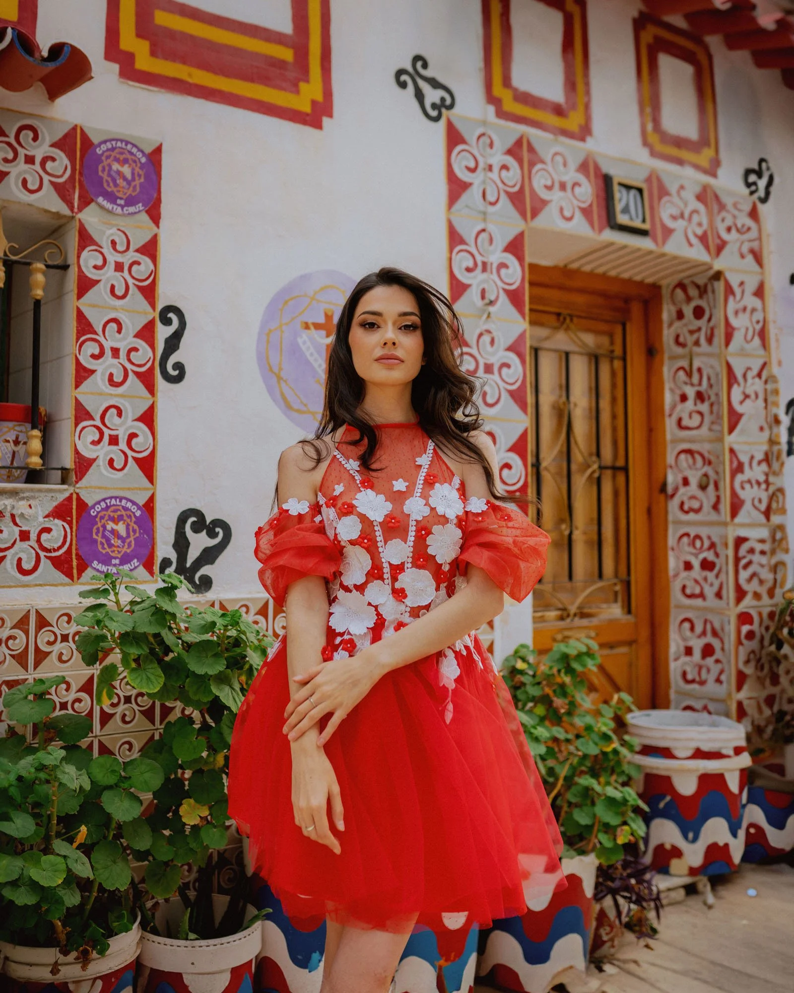 A woman in a red dress with white floral appliqué standing outdoors in front of a colorful, decorated wall with plants in pots.