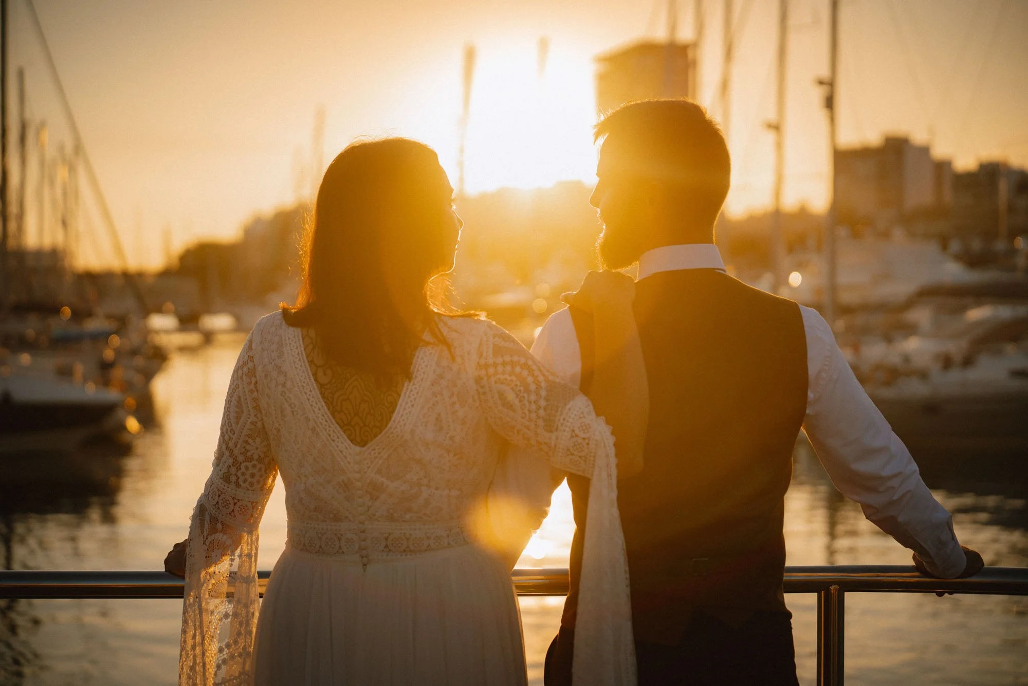 A couple standing on a boat at sunset, looking into each other's eyes with a marina and boats in the background. Alicante
