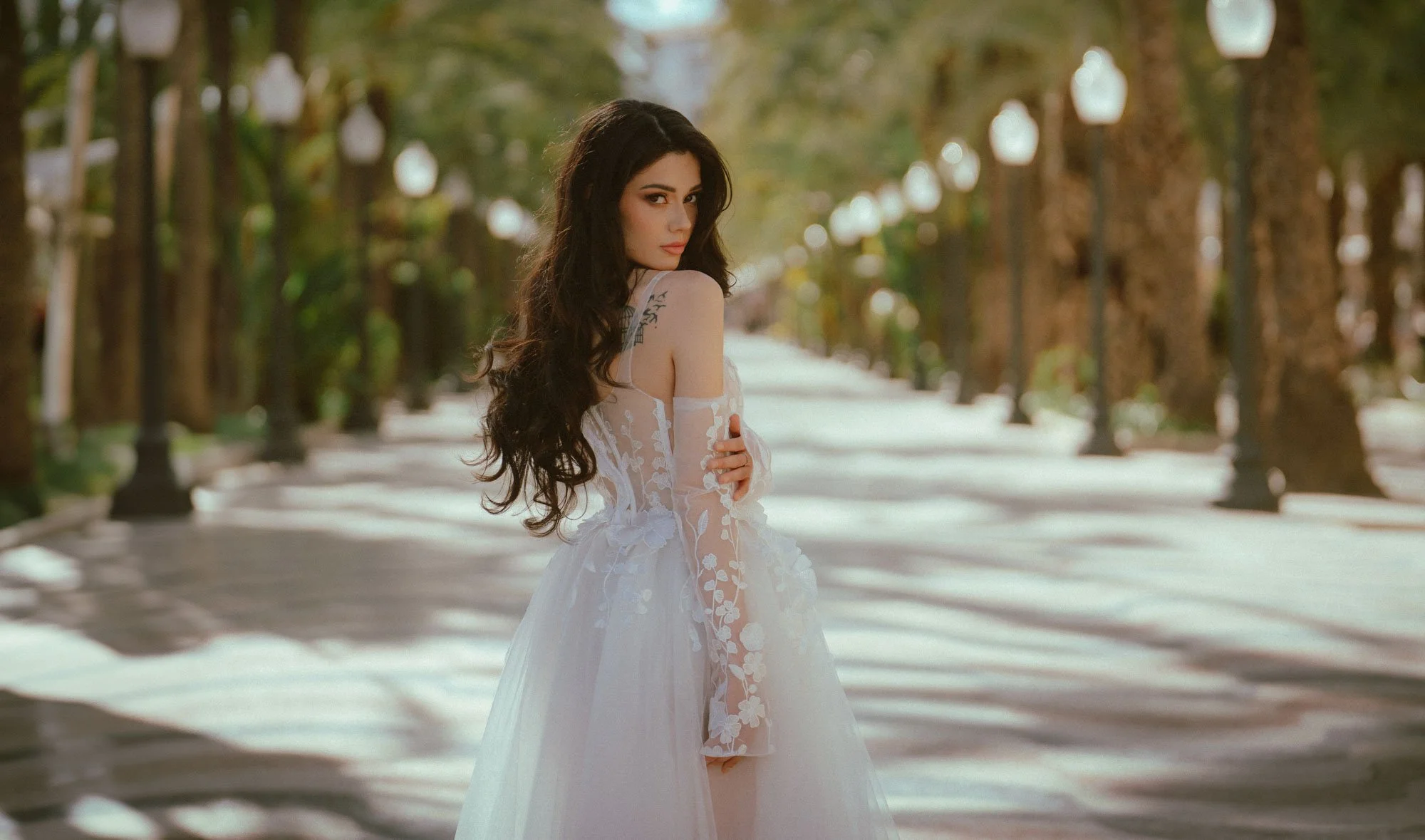 A woman with long dark hair in a white lace dress standing in a tree-lined park at sunset.
