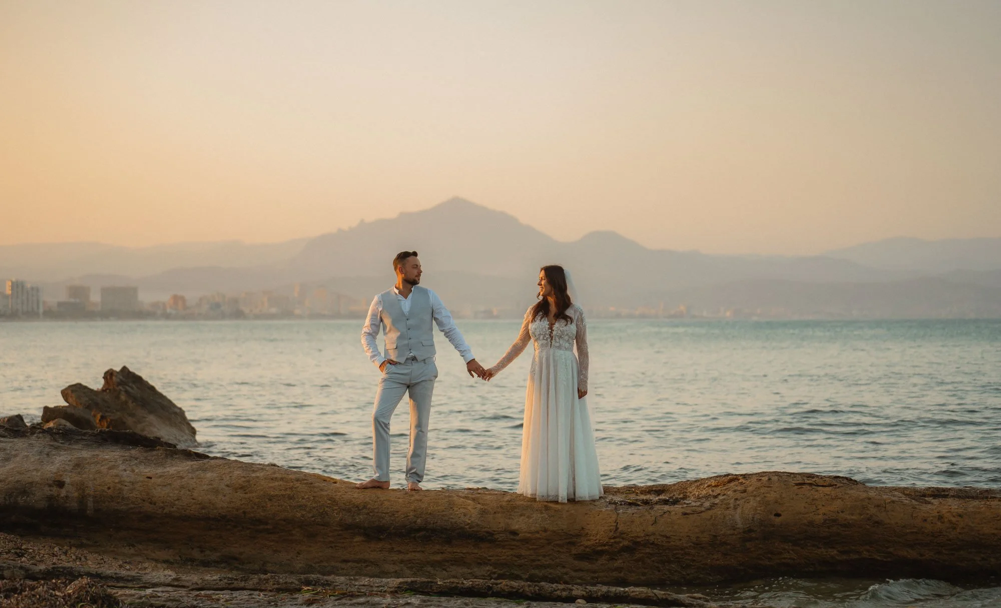 A couple in wedding attire holding hands on a rocky beach at sunset, with a city skyline and mountains in the background.