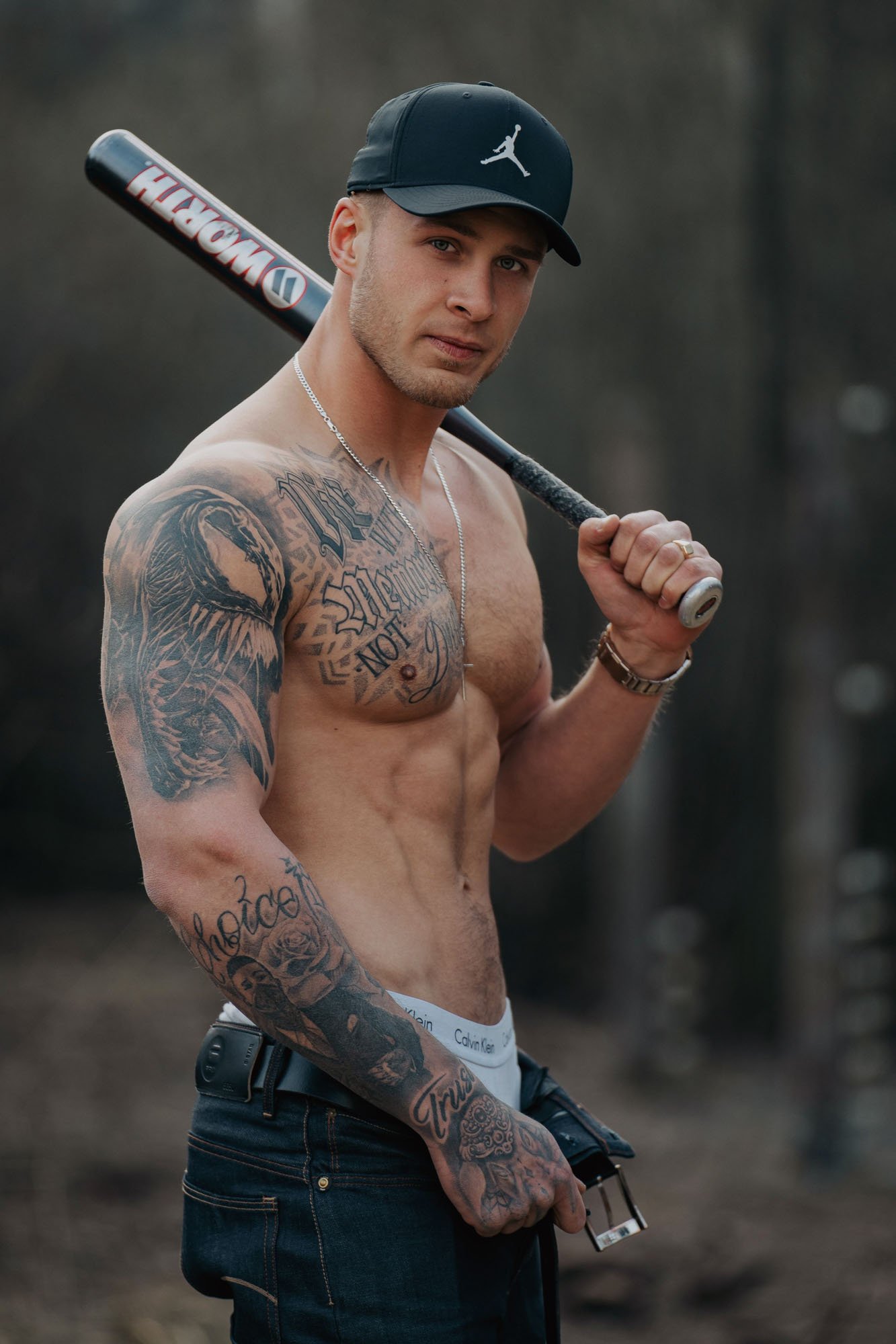 A shirtless young man wearing a black cap and jeans, holding a baseball bat over his shoulder, outdoors with a blurred background.
