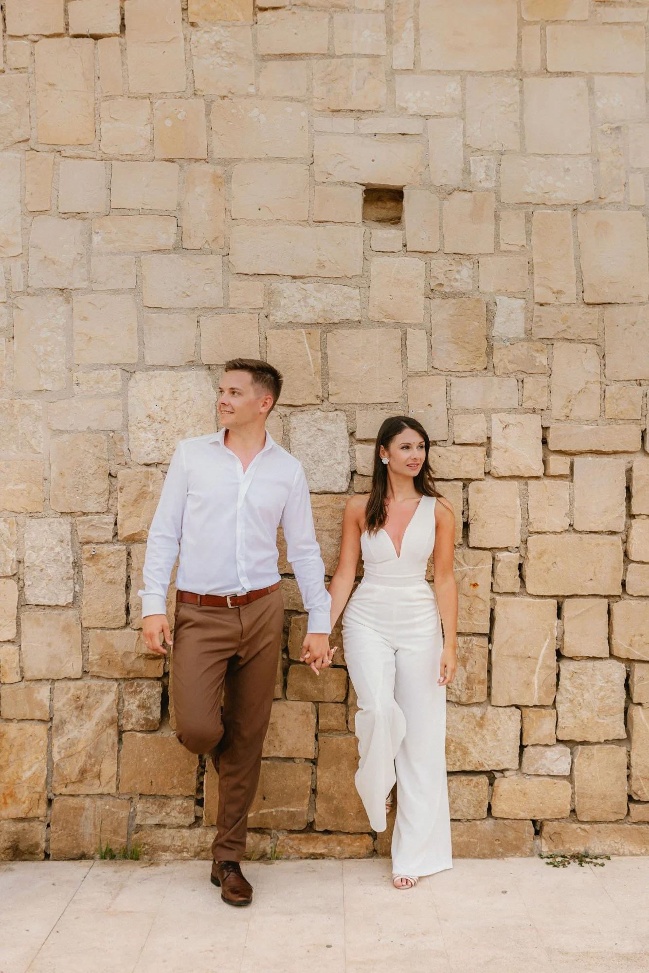 A man and woman holding hands and standing against a stone wall, dressed in formal attire.