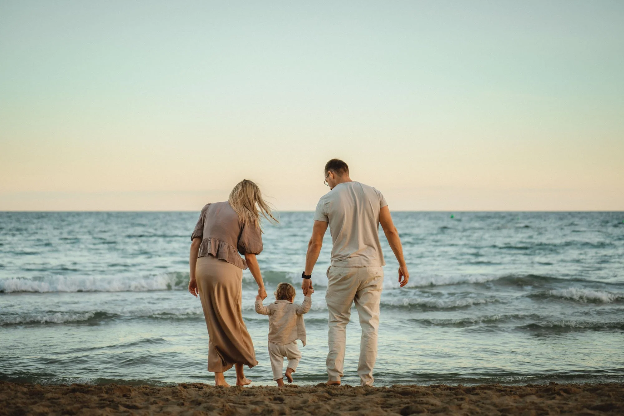 A family of three walking on the beach, holding hands, with the ocean in the background during sunset.