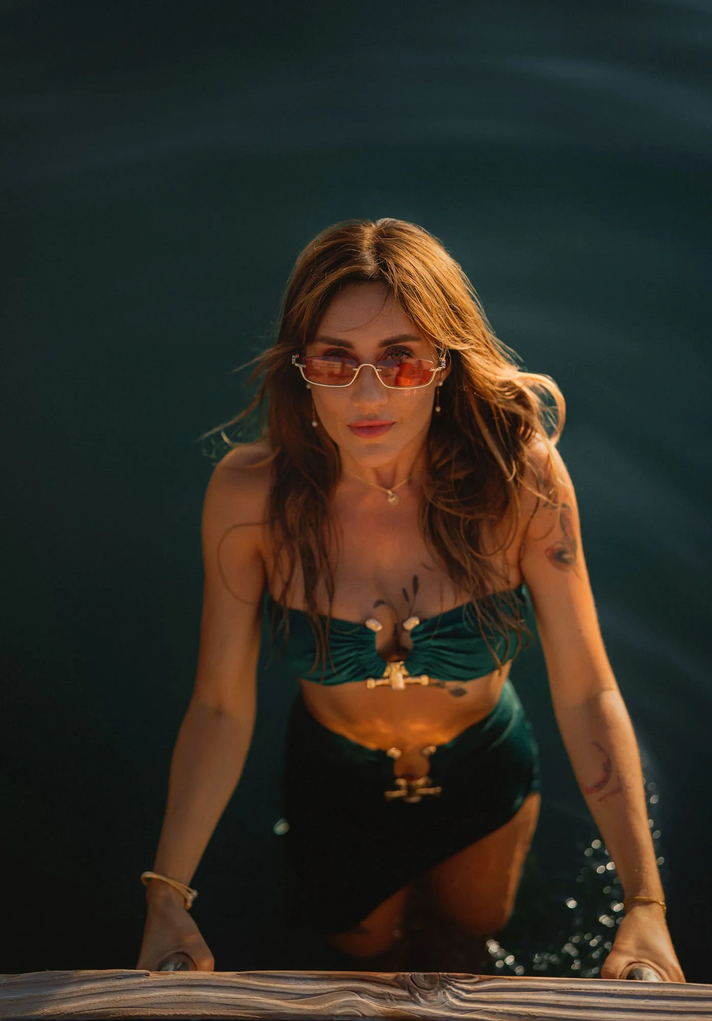 A woman with long brown hair wearing orange sunglasses, a green bikini top, and a matching green skirt, standing in water and looking up at the camera.