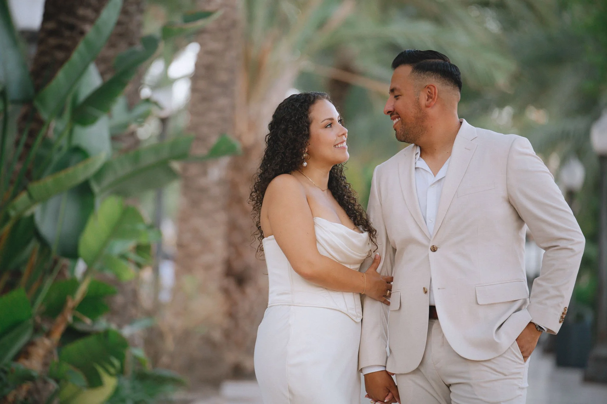 A couple in formal attire standing outdoors among palm trees, smiling and gazing at each other.