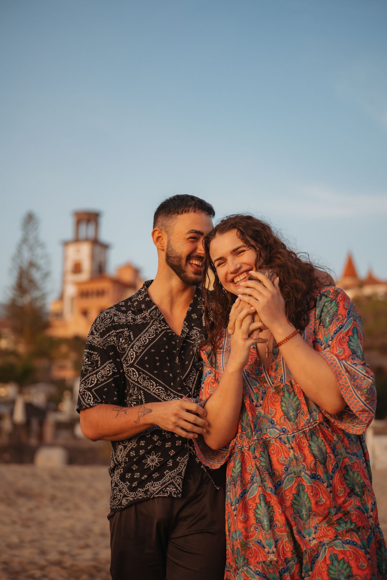 A joyful couple laughing and embracing on a beach at sunset, with a castle-like building in the background.
