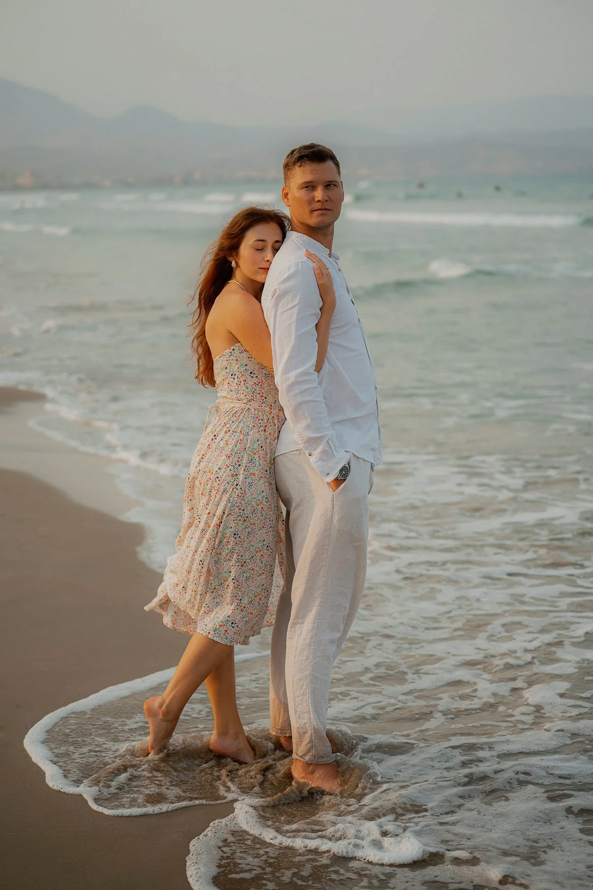 A couple stands close together at the edge of the ocean, with waves surrounding their feet. The woman leans on the man's shoulder, and both are dressed in light, summery clothing. The background features a distant shoreline and mountains under a cloudy sky.