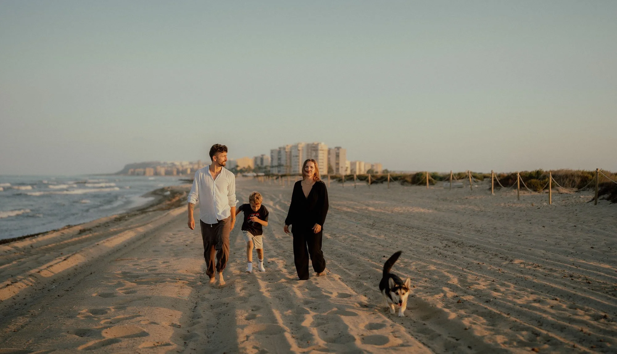 A family of three with a dog walking on a sandy beach at sunset, with buildings in the background.