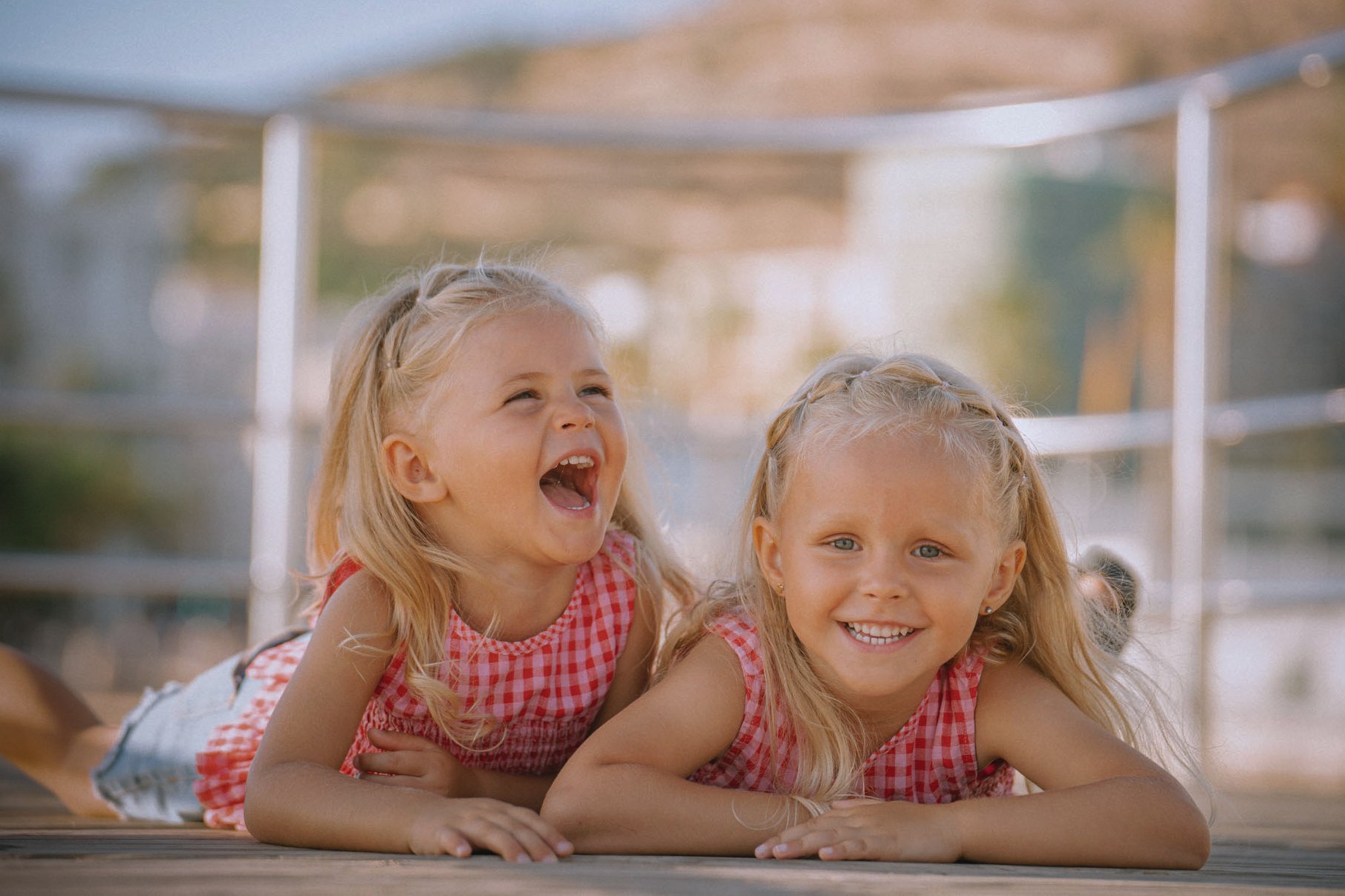 Two young girls with blonde hair lying on the ground outdoors, smiling and laughing, with a blurred outdoor background.