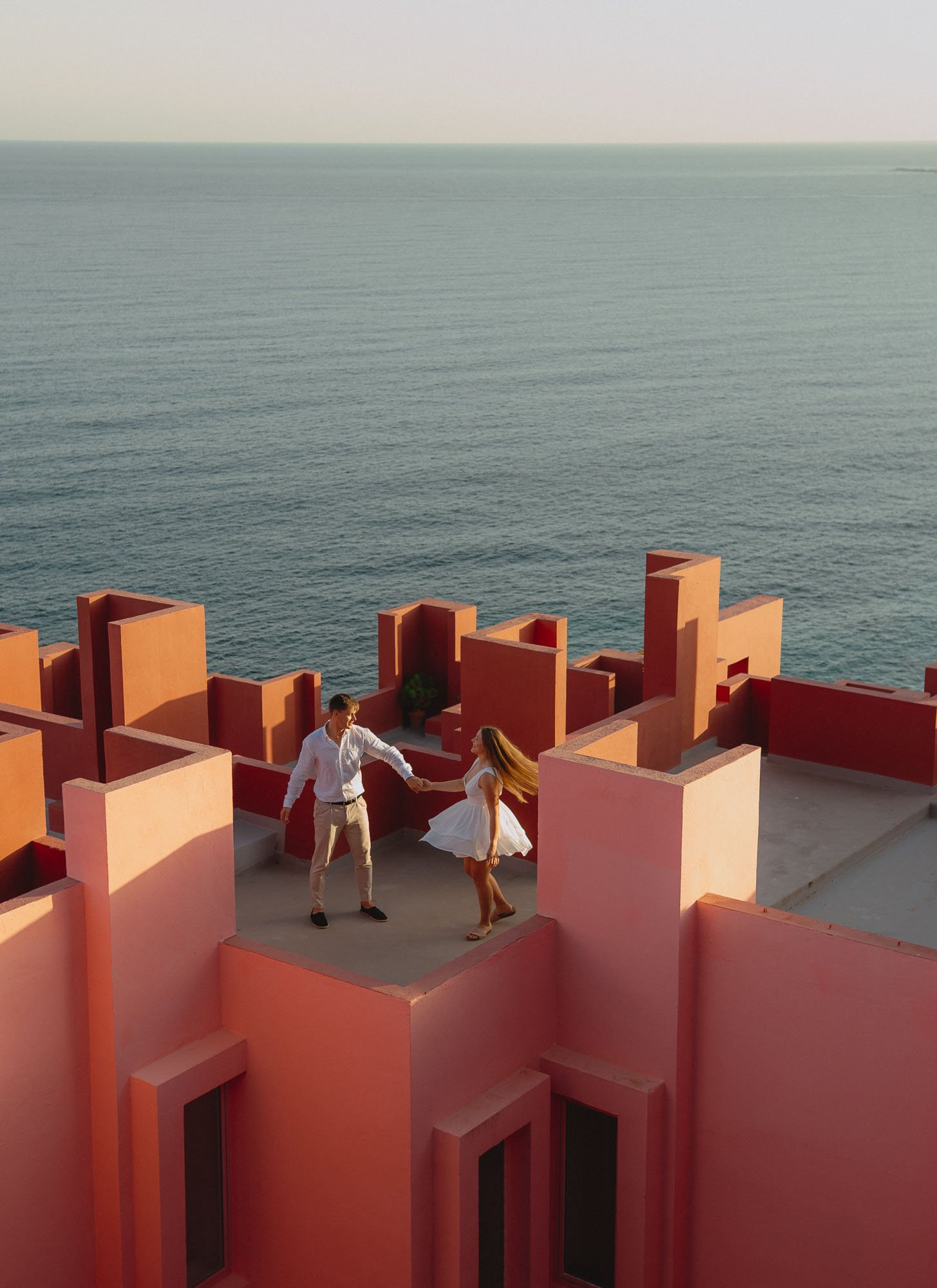 A young man and woman dancing on a pink rooftop near the ocean at sunset. Calpe, Muralla Roja Alicante