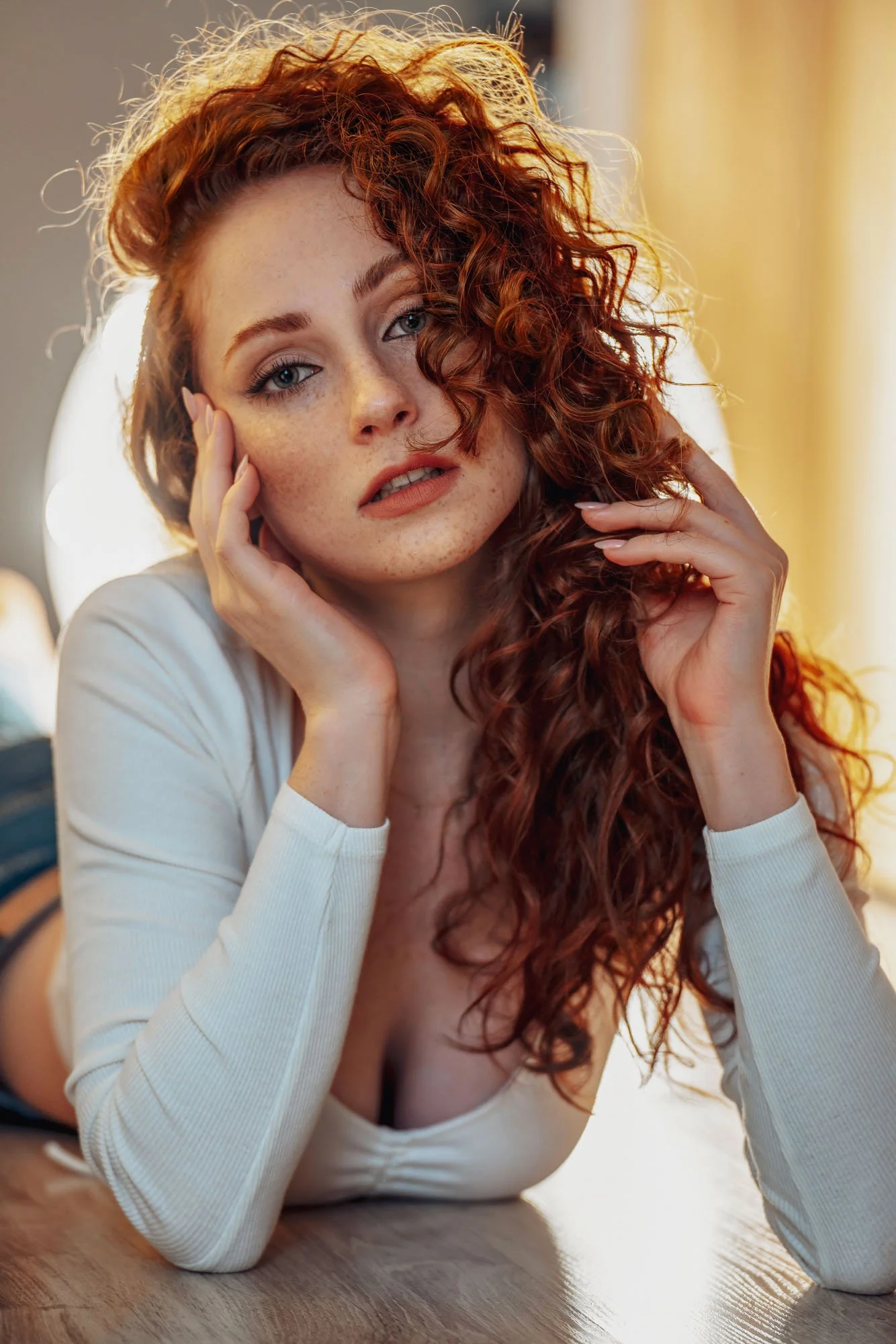 A young woman with red curly hair and freckles rests her face on her hand, gazing at the camera, indoors with warm lighting.