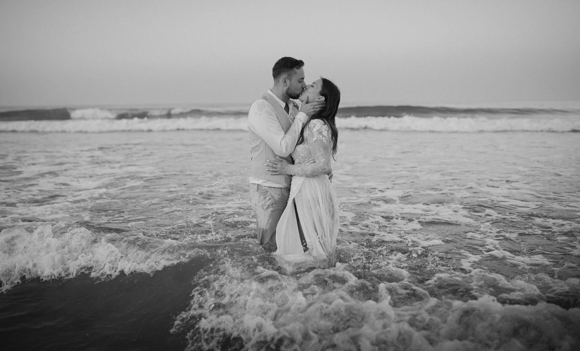 A couple in wedding attire kissing while standing in the ocean with waves around their legs.