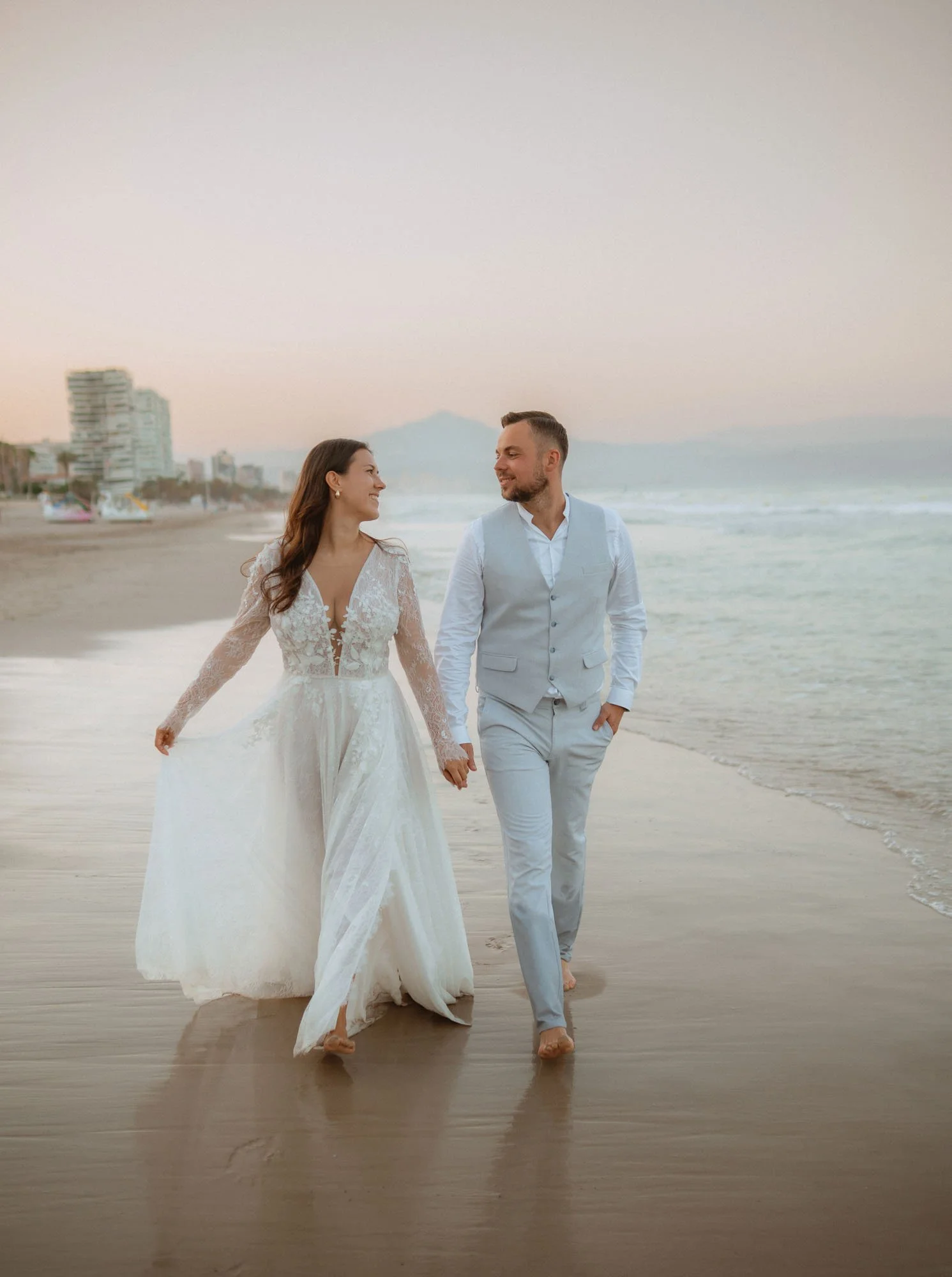 A bride and groom walk hand-in-hand on the beach during sunset, smiling at each other.