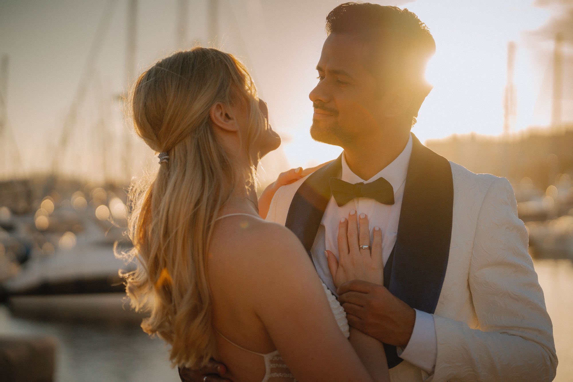 A bride and groom sharing an intimate moment outdoors during sunset near a marina, with sailboats in the background. Alicante
