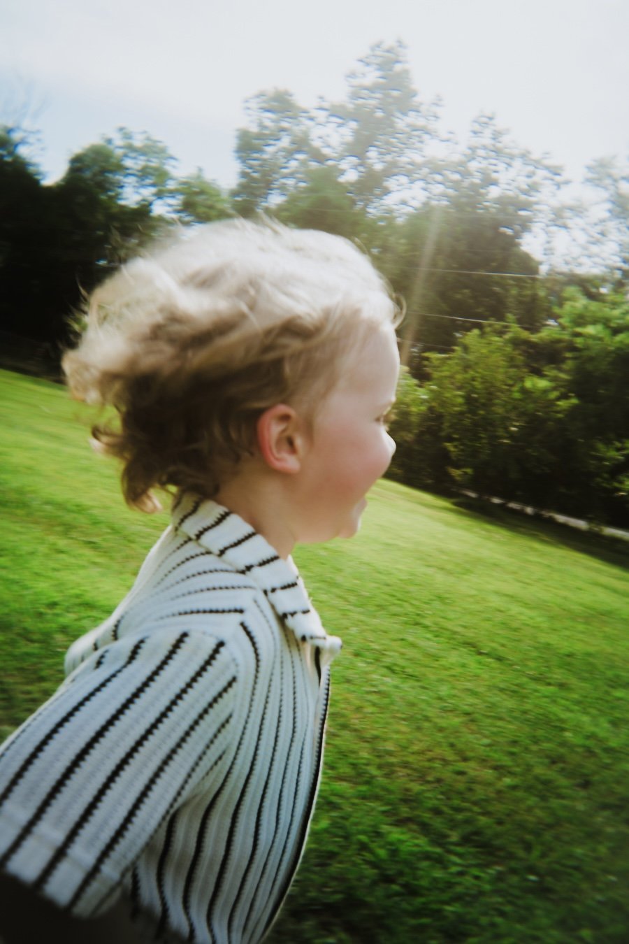 A young child with light brown, curly hair running outdoors on a green grassy field on a sunny day, wearing a white and black striped jacket.