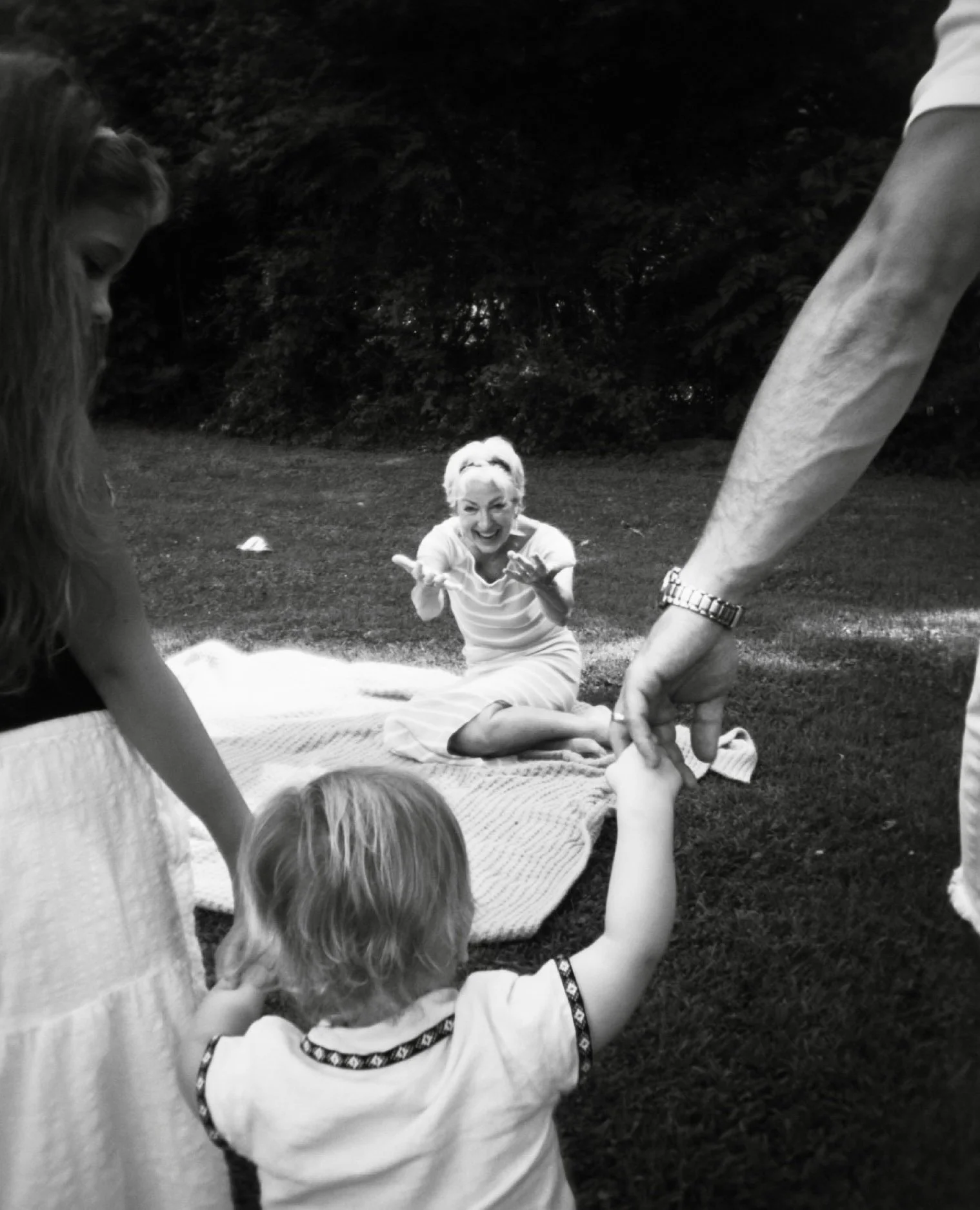A woman sitting on a blanket outdoors smiling, with children interacting with her and an adult holding a child's hand.